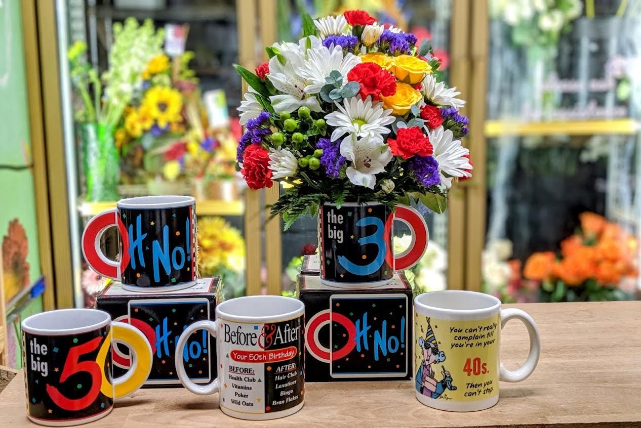 Colorful mixed flower bouquet in a mug-shaped container with novelty birthday mugs nearby