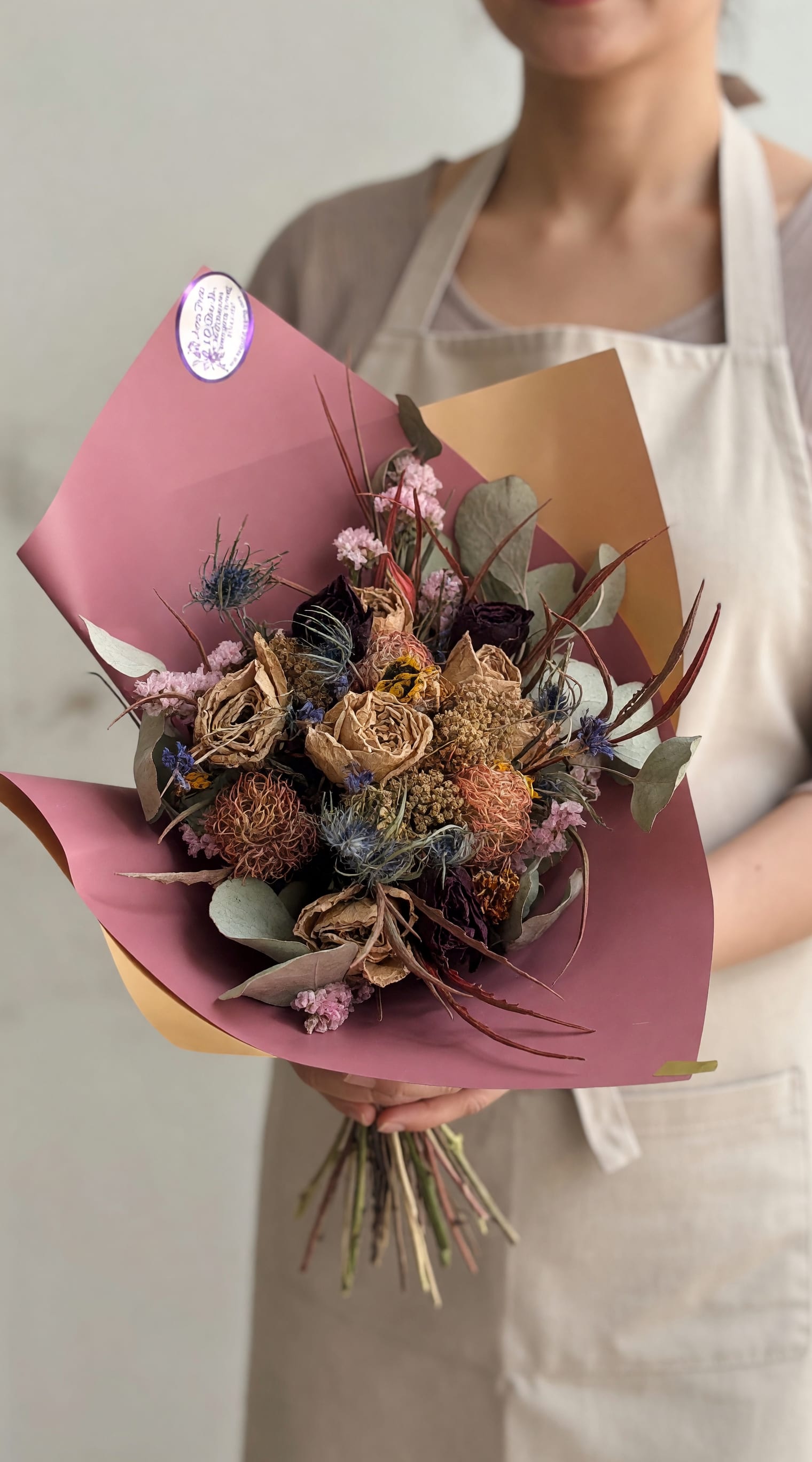 Vintage Blooms Dried Bouquet - Muted tan and dusty pink dried blooms arranged with deep purple and pale blue fillers give this hand-tied bouquet a soft, vintage look-perfect as an anniversary bouquet, a thoughtful "thinking of you" gift, or a long-lasting centerpiece for Thanksgiving. The compact, textured arrangement reads romantic and slightly rustic, ideal for someone who loves muted color palettes and heirloom-style decor.  Carefully wrapped in layered pink and warm beige decorative paper with a neat sticker label, the bouquet arrives ready to display without a vase. Because these are dried flowers, they keep their shape and color for months with minimal care-just dust gently and avoid direct sunlight. This makes it a charming choice for birthdays, new baby congratulations, get well wishes, thank yous, or simply to say "I love you" any time of year.