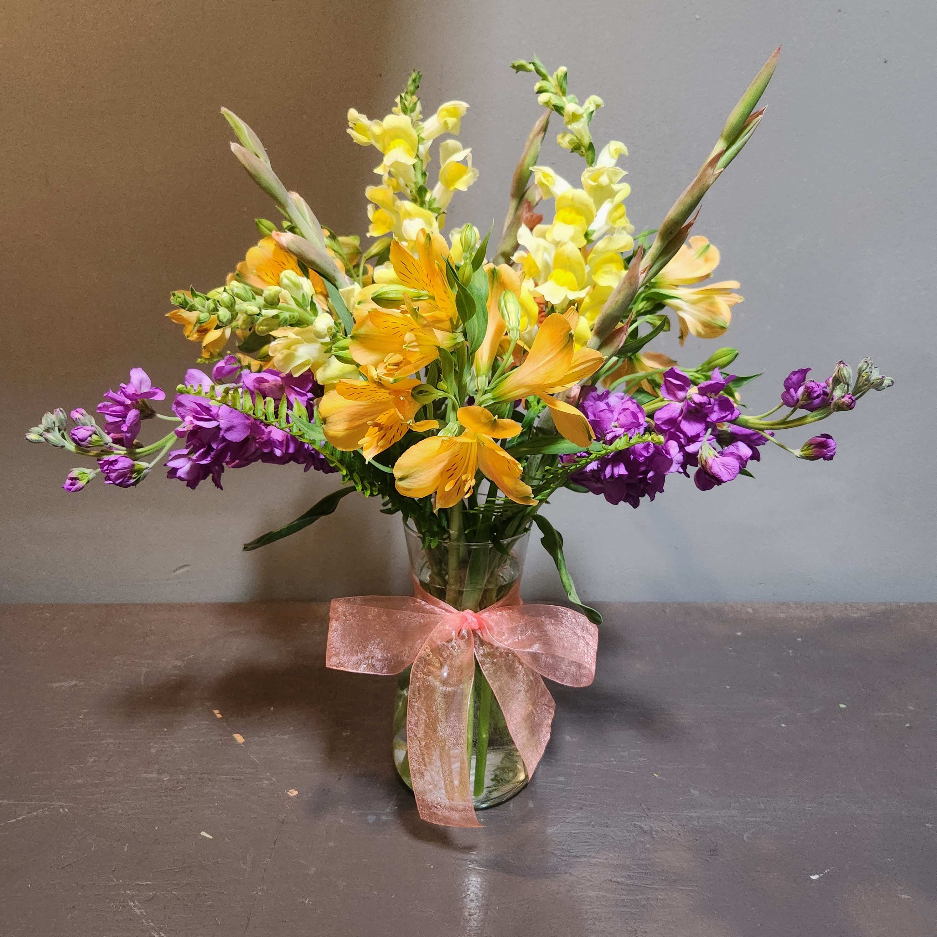 Mixed bouquet of yellow, orange, and purple flowers in a glass vase with a pink ribbon