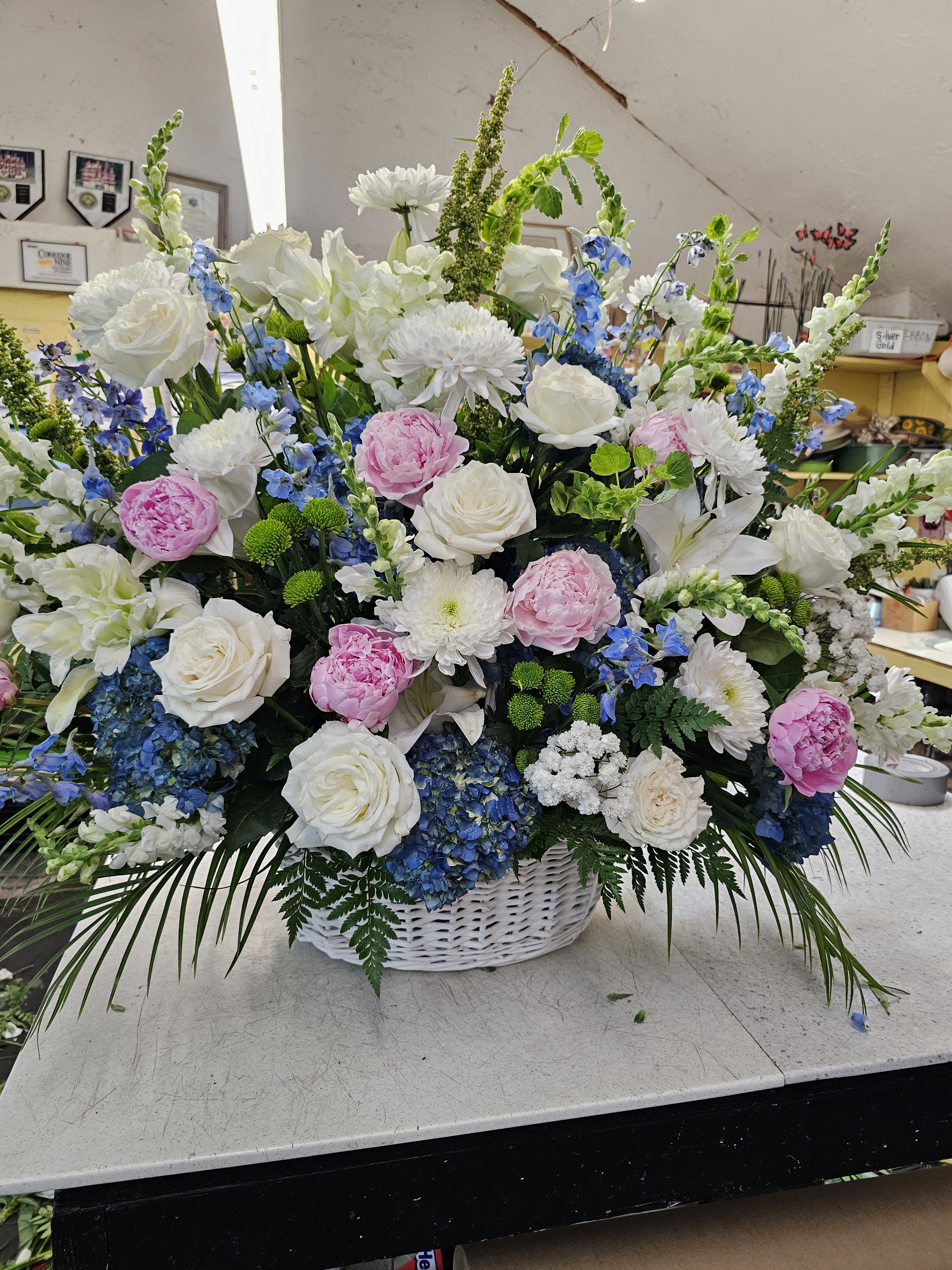 Faithful Memories Basket  - A sweep of soft blue hydrangeas and blushing pink peonies tops a cloud of white roses and chrysanthemums in this large, garden-style flower arrangement-perfect as a sympathy tribute, wedding centerpiece, or a heartfelt thank-you gift. The tall blue and white delphinium and snapdragon spikes add graceful height, so the person you're honoring will see a composed, layered design that reads both elegant and comforting from across the room.  Arranged in a classic white wicker basket with trailing greenery, the piece mixes round, lush blossoms with vertical accents for texture and movement. Expect abundant white roses, creamy chrysanthemums, pink peonies, blue hydrangeas, plus delphinium and snapdragon accents throughout; the basket presentation gives it a traditional, hand-tended feel that suits formal ceremonies or intimate home display. The overall palette of white, pink, blue, and green creates a serene, uplifting impression that helps the recipient feel remembered and cared for.  This is a substantial, full basket design meant to make an immediate visual statement-ideal for memorial services, bridal events, or to let someone know they're being held in your thoughts. Orders include the wicker basket and accompanying foliage; for best appearance, keep blooms in a cool spot away from direct sun and refresh the water if you separate stems into a vase.