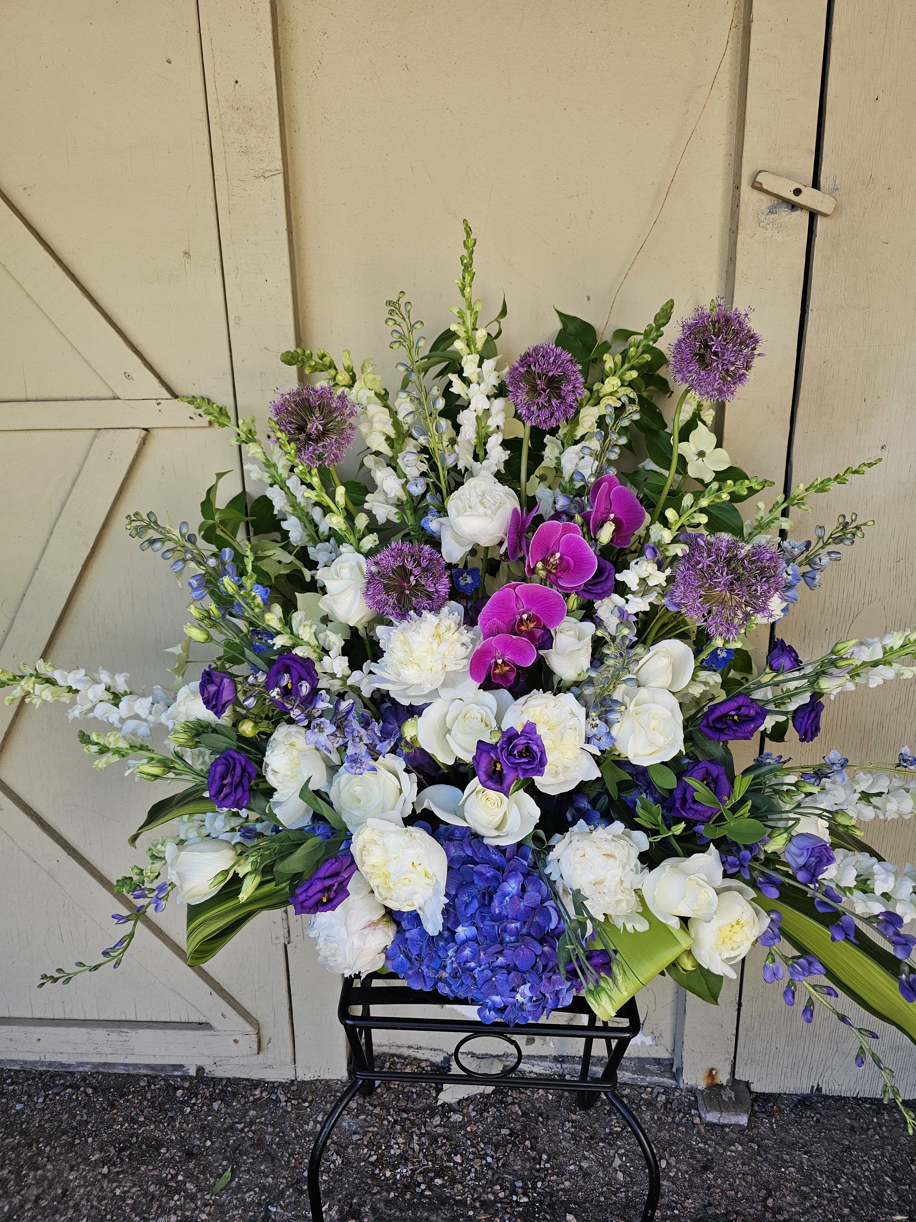 Royal Rememberance Tribute Basket - A dramatic, cool-toned sympathy arrangement crafted to honor a life with dignity and grace-the Royal Remembrance Tribute Basket reads as a formal memorial  designed for funerals or graveside services. The large upright silhouette  gives this funeral flower arrangement presence and height, so the person you're remembering is celebrated with a statement piece that feels both respectful and richly textured.  Dense white roses and hydrangeas form the luminous core, while vivid purple lisianthus and magenta orchids add elegant contrast and a touch of warmth. Blue accents and spherical purple allium blooms arc outward alongside tall snapdragons, creating long, layered lines that make the piece feel both full and graceful. The mix of whites, purples, blues, and greens produces a cool, refined palette suited to formal services.  