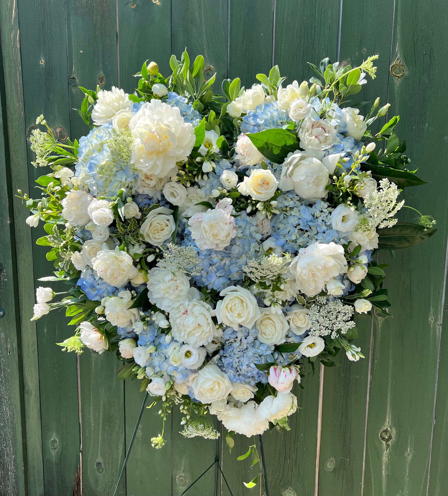 Full Heart - Honor a cherished life with this elegant White &amp; Blue Full Heart Easel, a timeless expression of love, remembrance, and heartfelt sympathy. Designed in the shape of a full heart, this beautiful standing arrangement features a graceful blend of pristine white roses, soft hydrangeas, lush peonies, and premium seasonal greenery, accented with rich blue blooms for a striking and comforting display. 