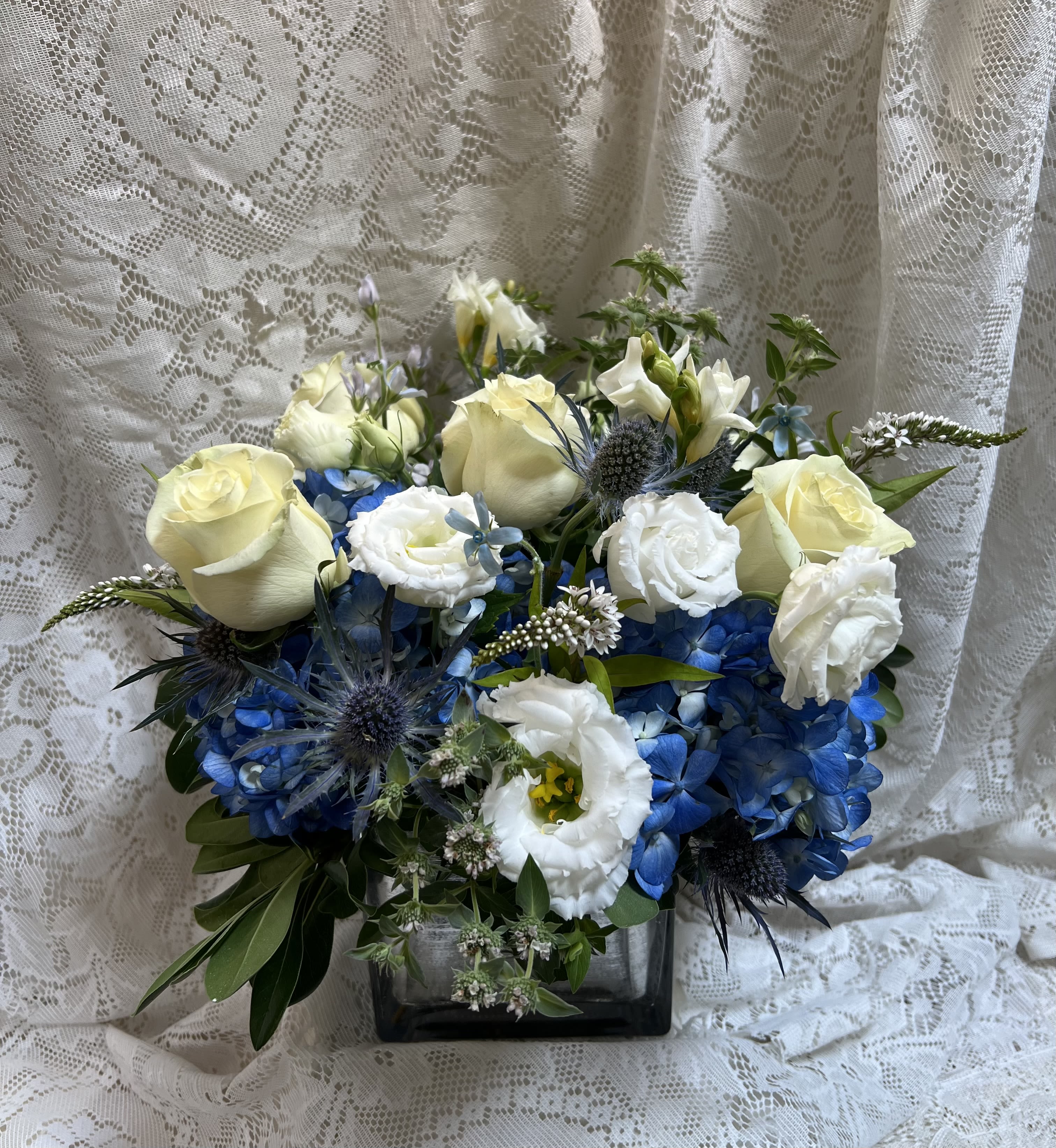 Skies of Blue - Soft, serene, and beautifully elegant, Skies of Blue captures the peaceful beauty of a clear blue sky. This arrangement features crisp white roses, delicate white lisianthus, textured blue thistle, and lush blue hydrangea, artfully designed in a modern low cube vase accented with a shimmering silver wrap.
