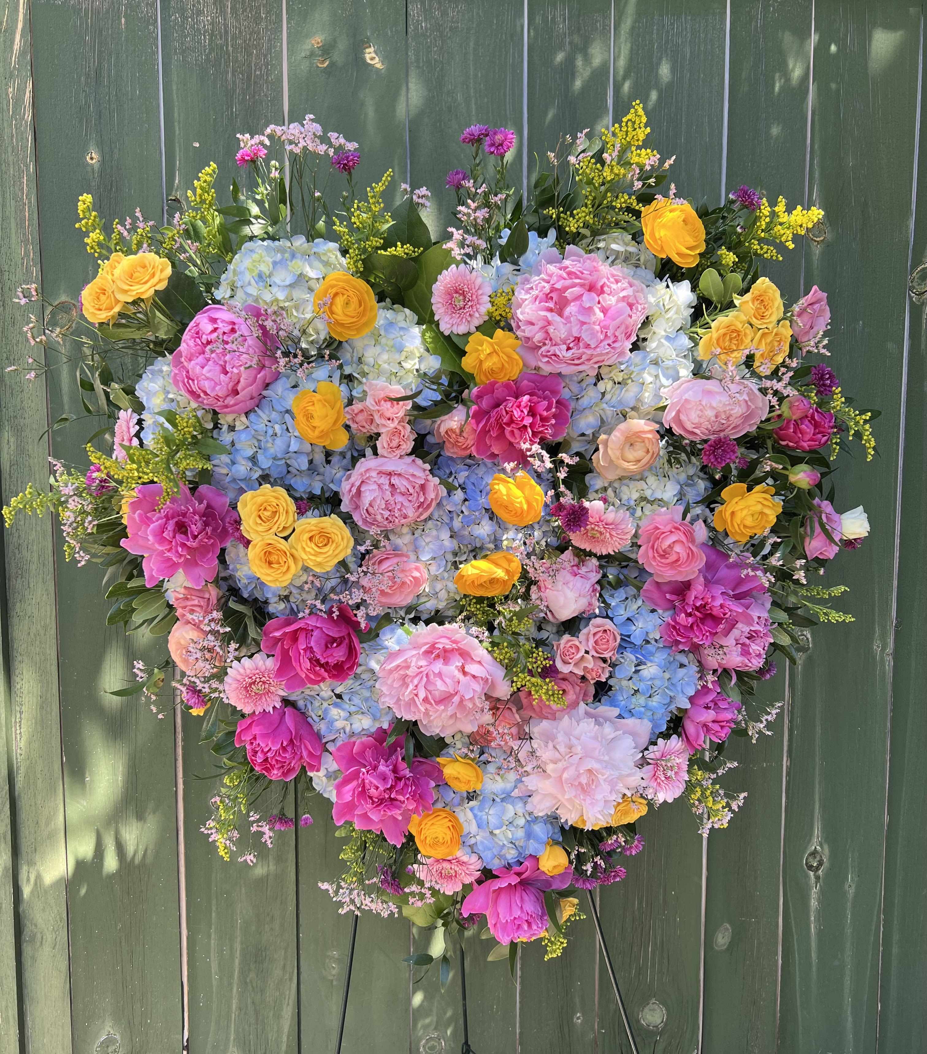 Heart of a Lifetime Tribute  - A lush, heart-shaped tribute overflowing with large peonies and ranunculus makes an immediate, romantic impression-perfect as a sympathy tribute or a statement piece for a memorial service. This flower arrangement heart combines pinks, cheerful yellows, soft blues and touches of lavender to create a warm, celebratory tone that honors a life while offering comfort to those who receive it.  Densely packed with layers of texture, the design pairs bold blue hydrangea clusters with small spray roses, daisy-like accents and other seasonal blooms to form a deeply dimensional surface. The colors read as both gentle and uplifting, and the mix of full peony heads and delicate ranunculus blossoms gives the piece a tactile, garden-fresh feel. The finished heart is presented on an easle highlighting its large scale and making it easy to display at a service or event.  For the person you want to remember, this arrangement says love, warmth, and celebration of a life well-lived-an especially fitting choice for memorials, funeral services, or a tribute at a celebration of life. 