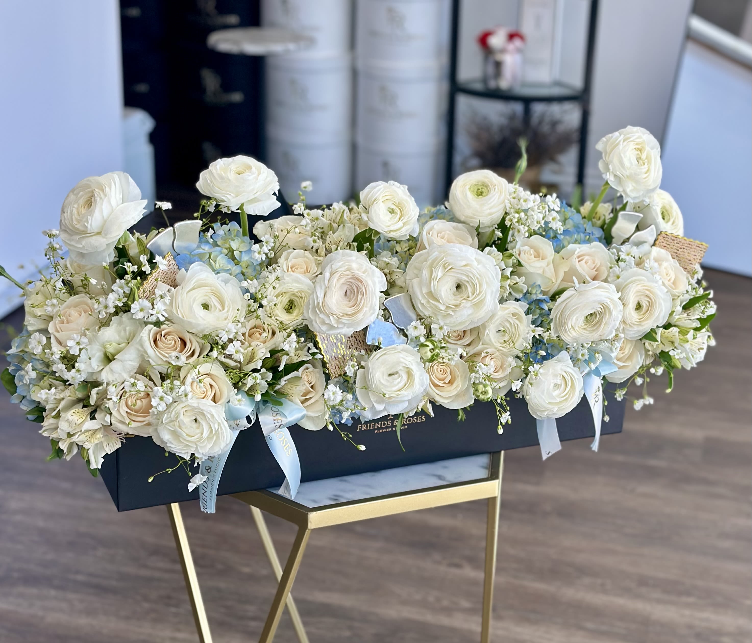 Traditions in Bloom - Celebrate Passover with timeless beauty and grace. This refined arrangement features soft ivory ranunculus, delicate white roses, and airy seasonal blooms accented with fresh greenery and subtle blue touches—evoking renewal, purity, and the spirit of the holiday. Designed in a modern keepsake box with elegant ribbon details, it makes a stunning centerpiece for your Seder table or a thoughtful gift to honor family and tradition.
