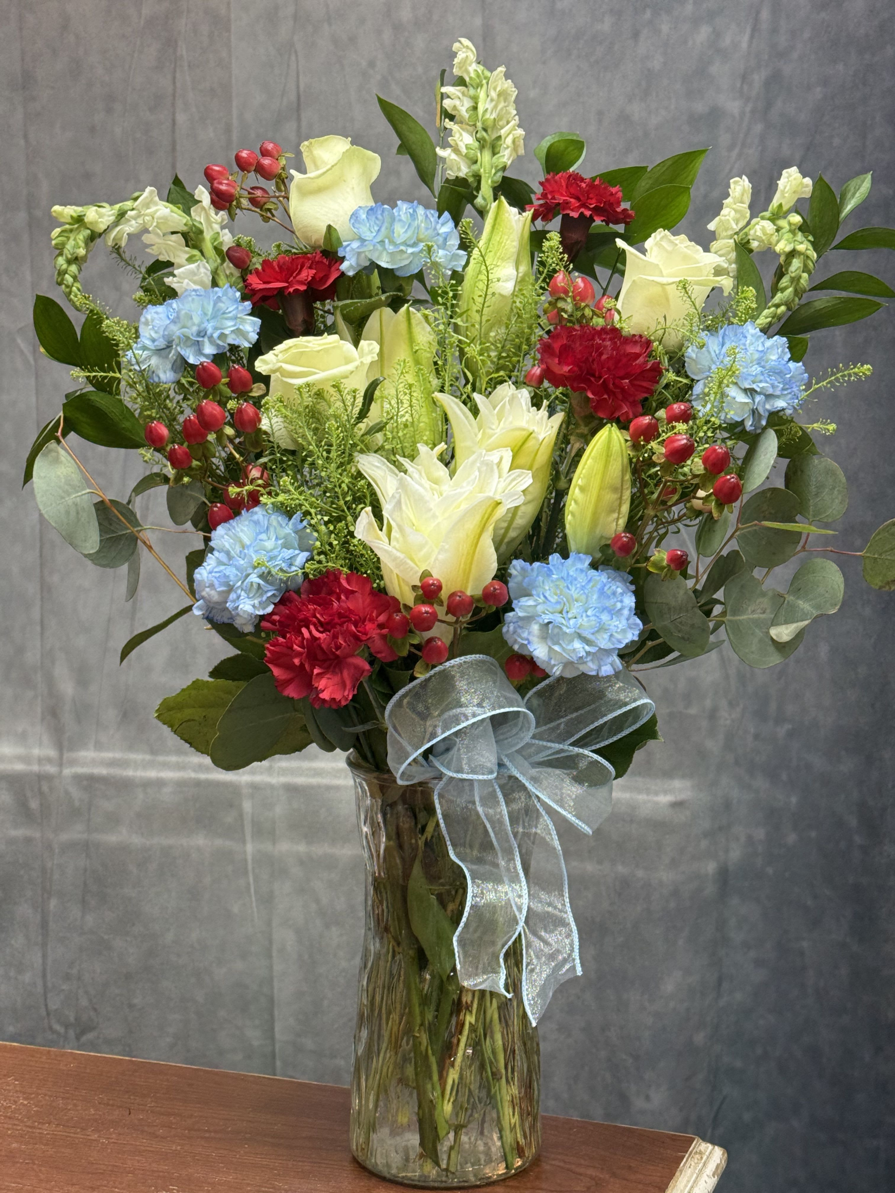 Lily of Honor Vase - A tall, full Lily of Honor vase arrangement that balances classic white lilies and roses with bright red carnations and soft blue blooms-perfect as a birthday bouquet, congratulations gift, or just-because surprise. The upright, abundant shape reads as both festive and slightly romantic, so the person you're thinking of will feel celebrated and genuinely seen.  This mixed-color arrangement sits in a clear glass vase and is finished with a sheer pale ribbon bow. Airy greenery, small red berry accents, snapdragons and other seasonal fillers add texture and movement around the edges, creating a polished but natural look that works for everyday gifting or more thoughtful occasions.  Arrives ready to display; vase included. Its generous height and full silhouette make it a striking centerpiece on a table or console, and simple care-fresh water and a clean vase-will keep blooms looking their best.