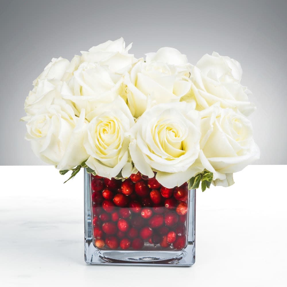 Compact arrangement of white roses in a clear glass cube vase filled with red berries