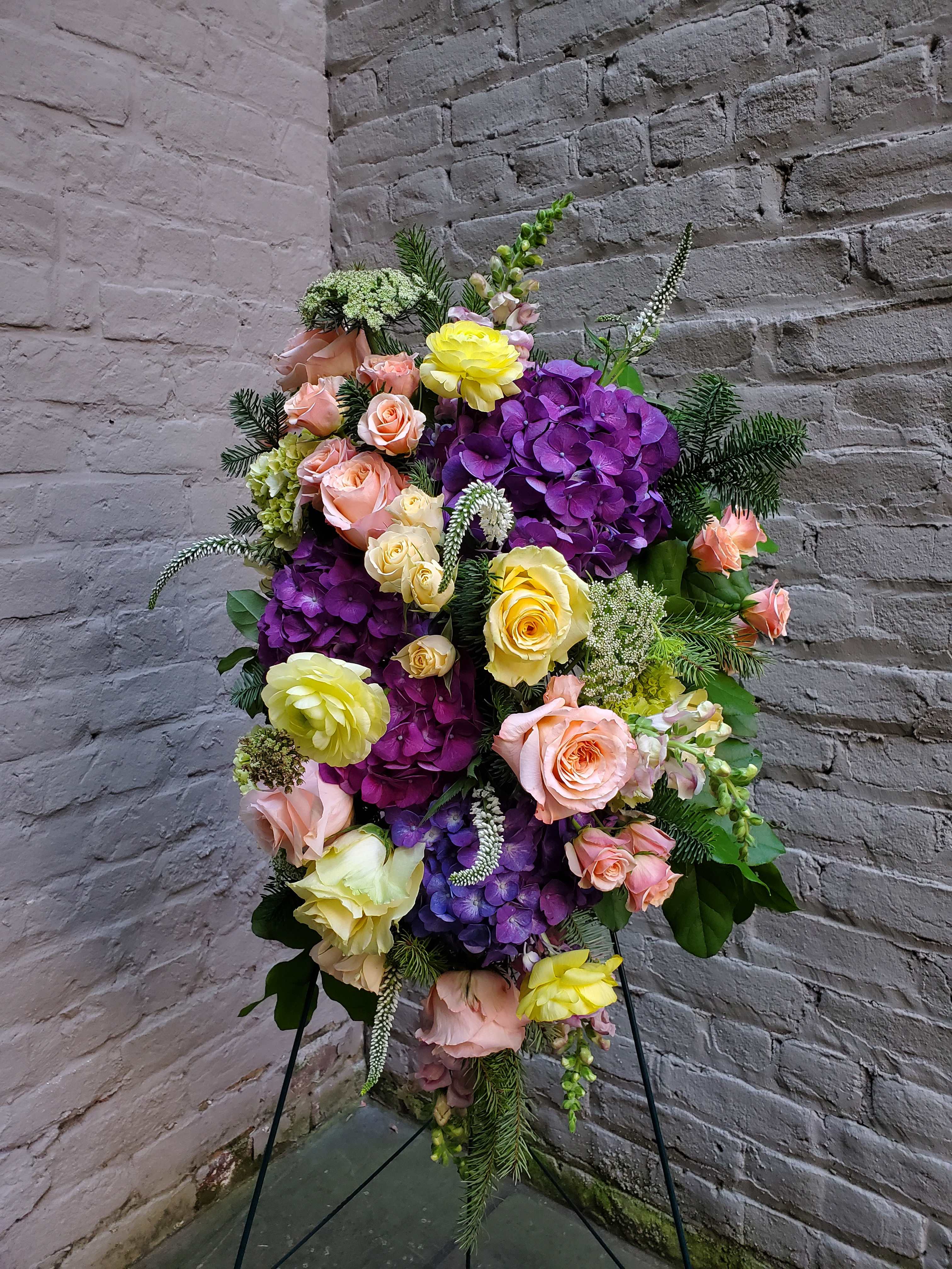 Standing Funeral Spray with Easel - A standing display of flowers to celebrate the life of your loved one.  Our photo features colorful blooms; it is our honor to accommodate any other specific color preferences, per your request. Delivered on wire easel. Measures approximately 22" long by 15" wide.  A special note: This arrangement is constructed with compostable basalt wool (in lieu of the typical floral foam and plastic). When available, we are happy to incorporate locally-farmed and harvested flowers as well. Thank you for supporting a more eco-friendly and sustainable option for funeral florals.