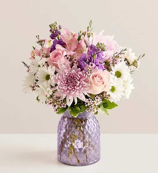 Pastel bouquet of pink roses, mums, daisies and purple blooms in a lavender glass vase