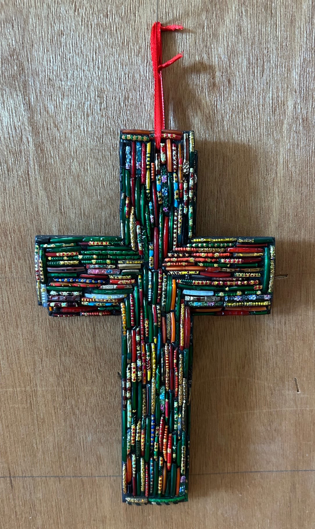 Colorful beaded mosaic cross ornament with a red hanging ribbon on a wood surface