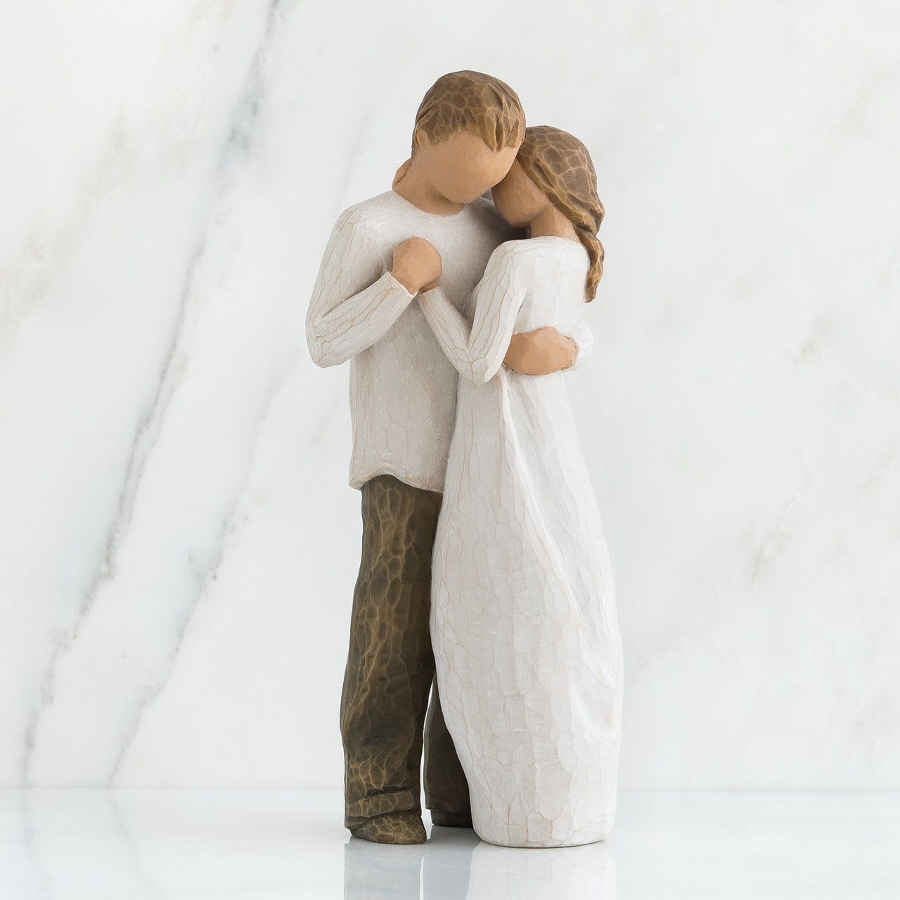 Promise - A quiet, intimate keepsake that speaks of lasting devotion-this Promise flower arrangement pairs a simple carved wooden figurine of a couple in an embrace with soft white blooms for a romantic anniversary or wedding gift. The couple's faceless, hand-finished look and neutral palette let the sentiment take center stage, making this the kind of arrangement that will make the person you're thinking of feel truly cherished and understood.  The arrangement features predominantly white flowers (clean, calming tones) arranged around the wooden figurine so the sculpture remains the focal point. The carved figures are painted in soft white and natural wood tones with a textured finish, offering a minimalist, decorative accent they can display long after the flowers have faded. Sized to sit on a tabletop or mantel, this piece combines living blooms with a lasting memento for a meaningful presentation.  Perfect for anniversaries, weddings, or any moment when you want to promise ongoing love and support, Promise is an understated, romantic choice for the person who appreciates both floral beauty and thoughtful keepsakes. For best enjoyment, keep the flowers in a cool spot away from direct sunlight and change the vase water daily to prolong the life of the blooms; the wooden figurine requires no care beyond gentle dusting.