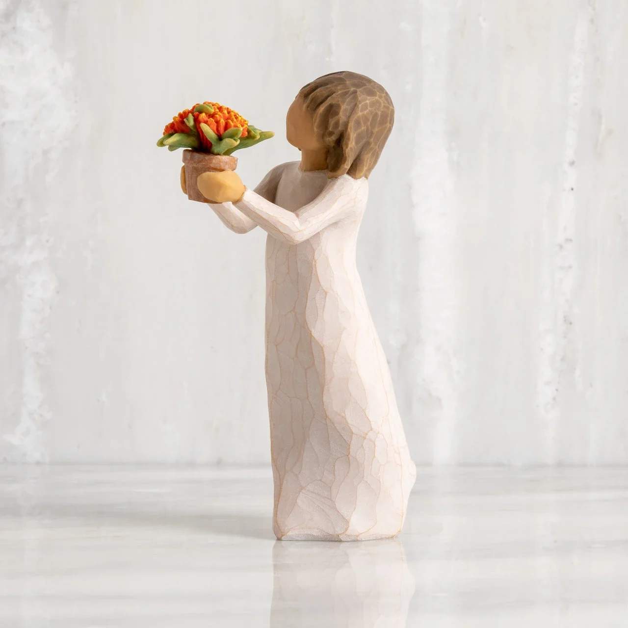 Willow Tree Little Things - A sweet, sentiment-forward gift that looks like a keepsake as much as a floral piece - this figurine arrangement is perfect as a sympathy gift, a "thinking of you" surprise, or a small anniversary token. The decorative girl in a long white dress cradles a tiny potted bouquet, so the recipient will feel quietly remembered when they see the gentle, handcrafted scene.  The compact faux bouquet features bright orange stylized blooms with fresh green accents set in a small brown pot, contrasting softly against the neutral, matte finish of the figure. Because this is a hard-good figurine rather than fresh flowers, it stays beautiful without water or maintenance and works well displayed on a bedside table, mantel, or office desk.  Ideal for someone who treasures meaningful mementos, this piece sends a calm, thoughtful message - the kind that comforts and endures. Sized to fit into small spaces, it arrives ready to display and requires no care, making it an easy, lasting way to show you care.