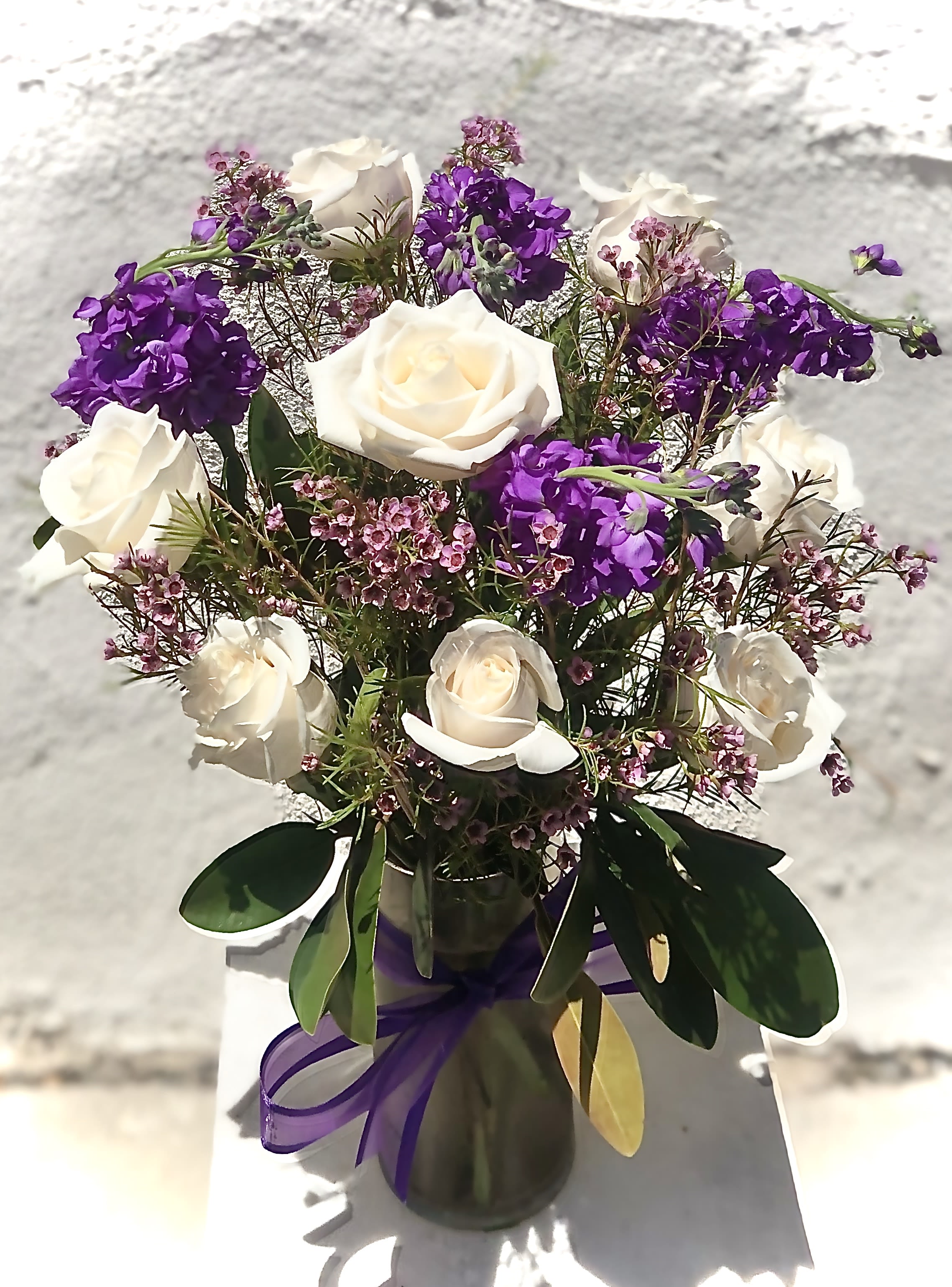 White roses and purple flowers arranged in a glass vase with ribbon