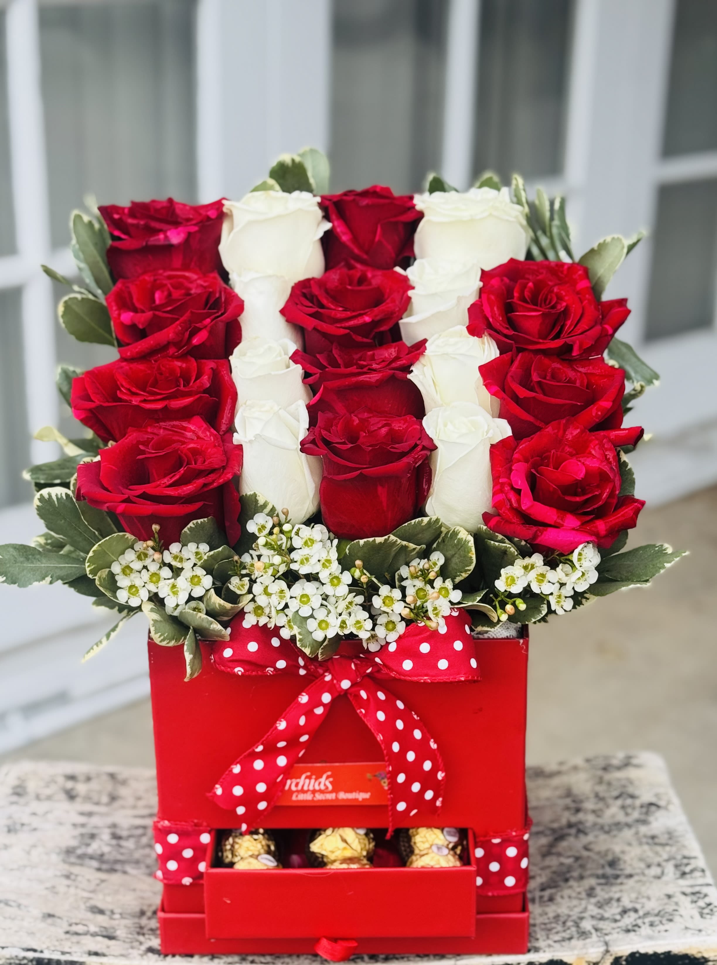 Red and white roses arranged in a red gift box with chocolates and a polka-dot ribbon