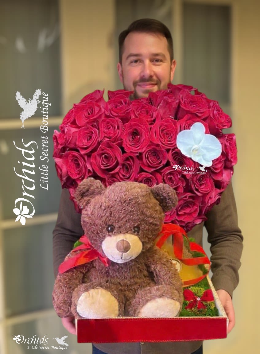 Man holding a heart-shaped rose arrangement with a teddy bear and ribbon