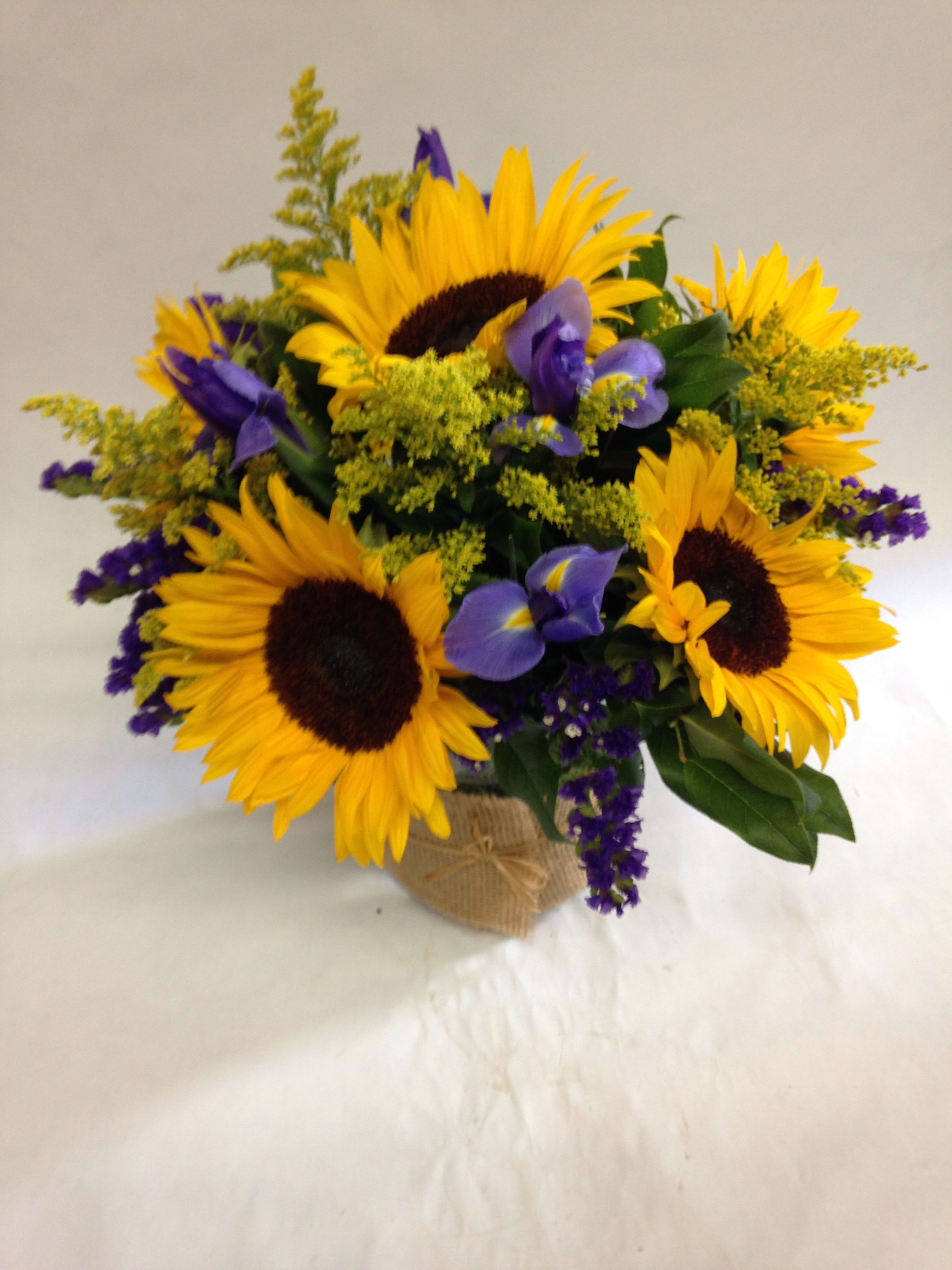 Arrangement of bright yellow sunflowers with purple irises in a glass vase wrapped in burlap.