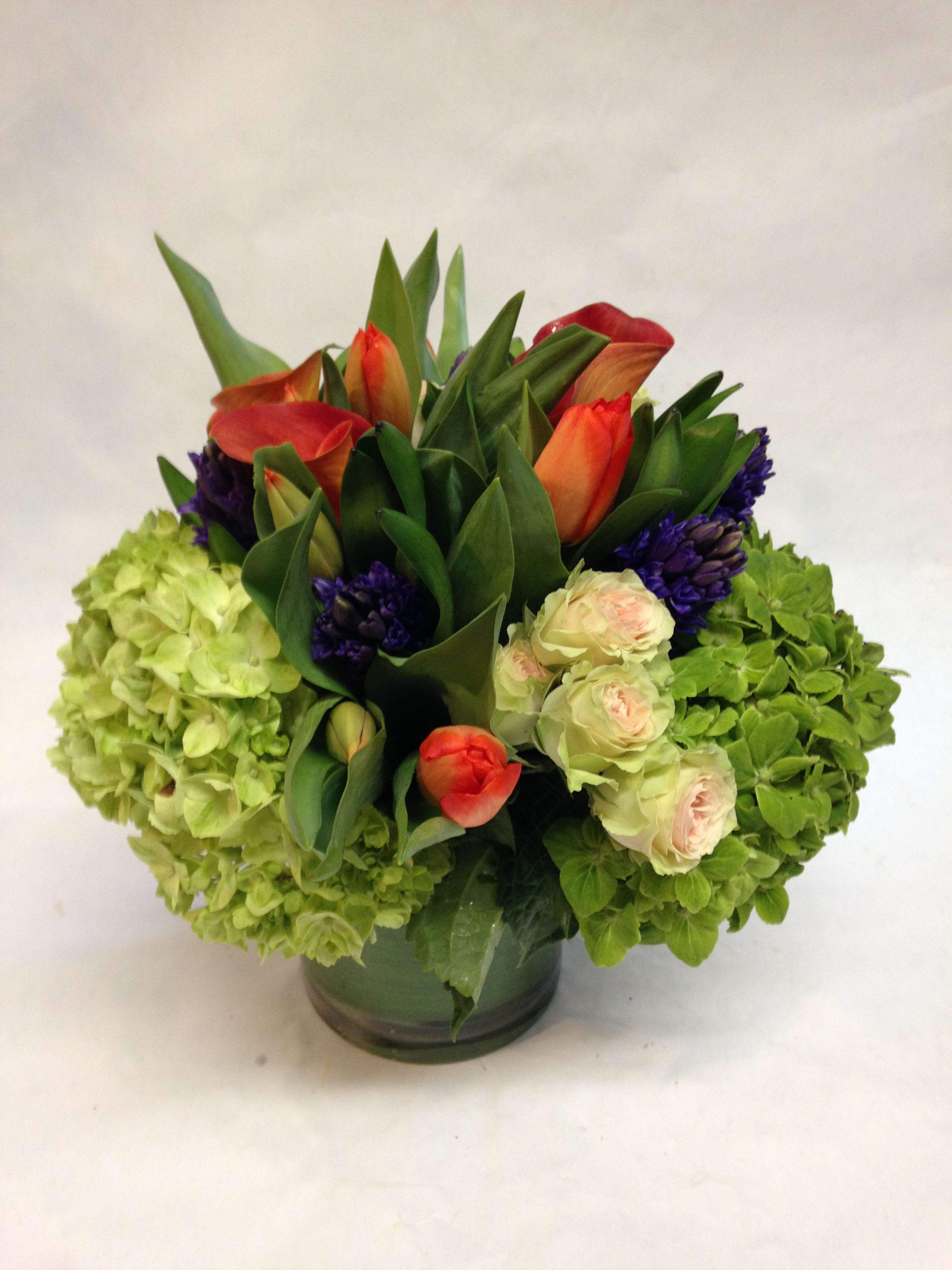Compact arrangement of green hydrangeas, orange tulips, cream roses and purple blooms in a round glass vase