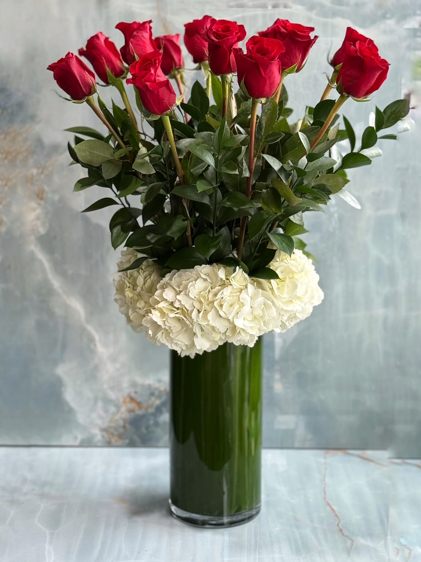 Red roses arranged in a tall glass vase with white hydrangeas at the base.