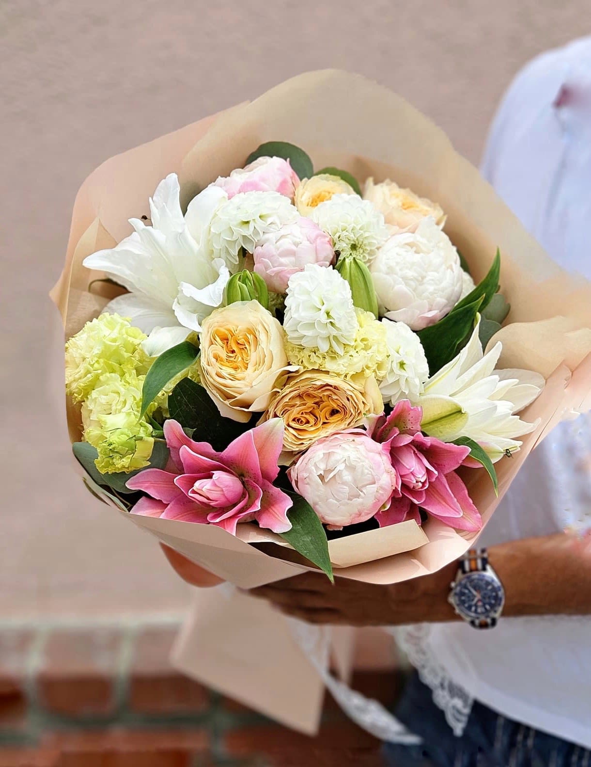 Hand-tied bouquet of white and pink lilies, roses, peonies, and mixed blooms wrapped in beige paper.