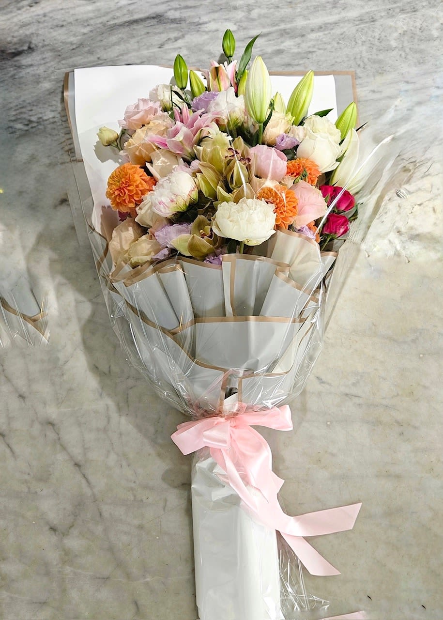 Soft Oyster Meadow - This bouquet is a soft, romantic mix of garden-inspired blooms in delicate pastel tones. Creamy whites, blush pinks, peach, and hints of lavender are layered together with elegant lily buds, ruffled roses, peony-style blooms, and cheerful pops of orange for warmth. Fresh greenery and unopened buds add movement and just-picked feel. Finished in crisp white paper and tied with a pale pink ribbon, it feels airy, refined, and effortlessly beautiful beautiful-perfect for celebrations, thank-yous, or simply brightening someone's day. 
