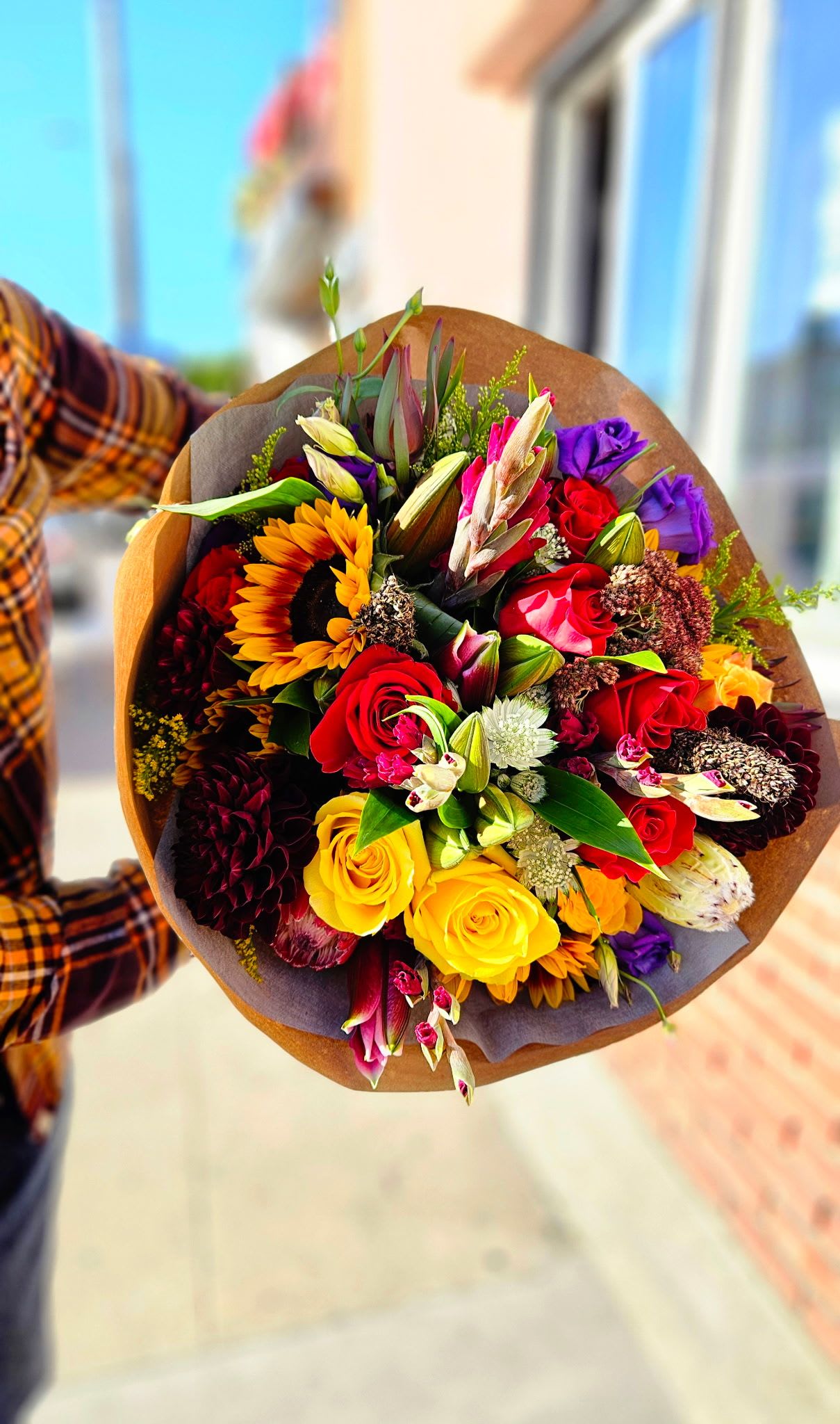Handheld bouquet of mixed roses, sunflowers, and lilies in brown wrap