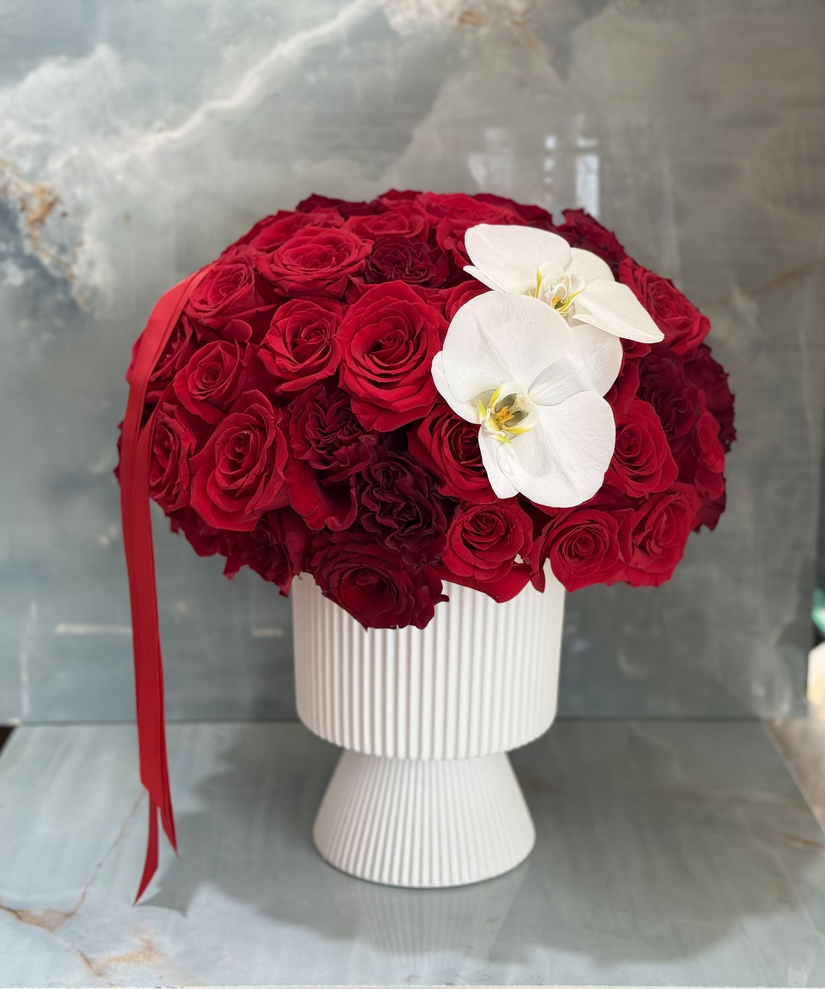 Red rose arrangement in a white pedestal vase with two white orchids