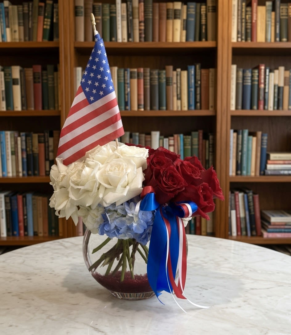 God Bless The USA - This arrangement is a graceful tribute to timeless American elegance. Rich red roses, pristine white roses, and soft blue hydrangeas are thoughtfully composed in a clear glass vase, accented with a patriotic ribbon and American flag. The balanced palette and classic design honor both beauty and country with dignity and sophistication. 