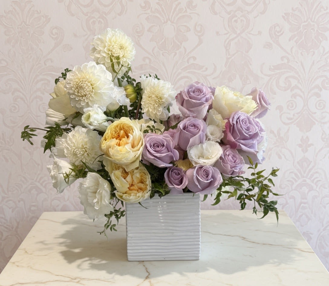 Rosé Champagne - Lavender roses and creamy white blooms mingle in a soft, pastel presentation that feels like a gentle toast-perfect as a birthday bouquet, anniversary arrangement, or a thoughtful thank-you gift. The Rosé Champagne flower arrangement brings a calm, elegant look that will make the person you're thinking of feel cherished and seen, whether for a spring celebration or just because. This compact, rounded design pairs soft purple roses with pale pink and yellow accents, creamy white lisianthus, and large white dahlia-like flowers that add fullness and texture. The blooms are arranged tightly for immediate impact and sit in a ribbed white square ceramic vase that gives the piece a clean, modern silhouette-easy to place on a mantel, desk, or dining table.