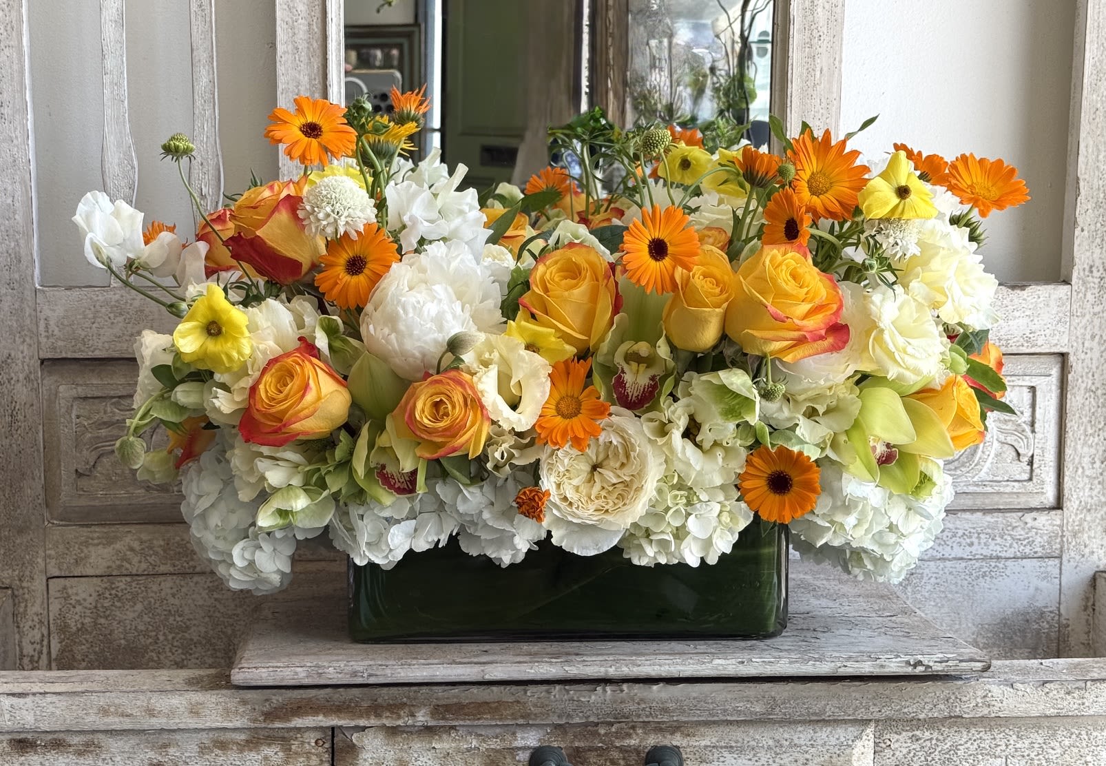 Summerfield  - A lively, sunlit mix that brightens any celebration-this mixed-color summer bouquet is a cheerful everyday arrangement perfect for birthdays, congratulations, or springtime get-togethers. Packed low and wide in a rectangular dark green glass vase, the composition presents a bold contrast between warm orange and yellow roses and large clusters of pure white hydrangeas, accented by bright gerbera daisies for a playful pop.  The flowers are closely arranged for a full, abundant look: soft-petaled roses in citrus hues, fluffy white hydrangea heads, and vibrant gerberas combine for varied texture and eye-catching color. The arrangement sits in a rectangular glass vase that keeps the design modern and compact, ideal for tabletops or office desks where the recipient will see it often.  Made to make someone feel celebrated and noticed, this bouquet says "congratulations" or "happy birthday" without being fussy. Its low profile makes it easy to place on a dining table or reception counter; simply keep the vase filled with fresh water and trim stems as needed to extend bloom life.