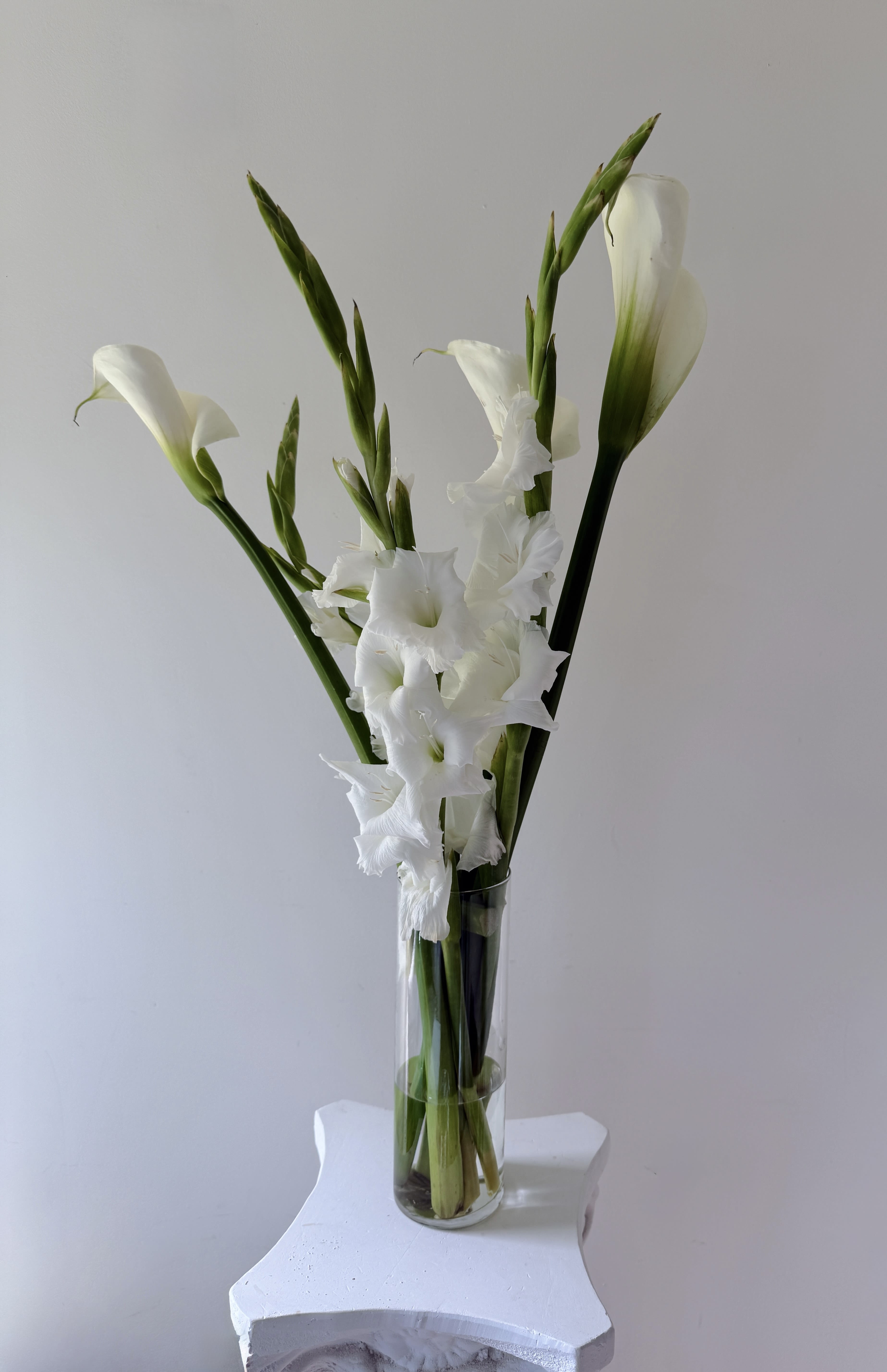 White Calla &amp; Gladiolus Arrangement - This floral design features white calla lilies and white gladiolus, two blooms known for their natural elegance and sculptural form.  The calla lilies bring a soft, curved silhouette with a clean and refined presence, often associated with minimal and modern floral design. Their simplicity creates a calm and sophisticated visual effect.  The gladiolus add height and structure, with tall stems and multiple blossoms that introduce vertical movement and balance to the arrangement.  Together, these flowers create a composition that feels light, modern, and effortlessly elegant, allowing each element to stand out without overwhelming the design.  Presented in a clear glass vase, the arrangement highlights the natural beauty of the stems and water, enhancing its fresh and authentic aesthetic.  Ideal for those who appreciate refined floral design, this piece brings a sense of calm, light, and understated luxury to any space or occasion.