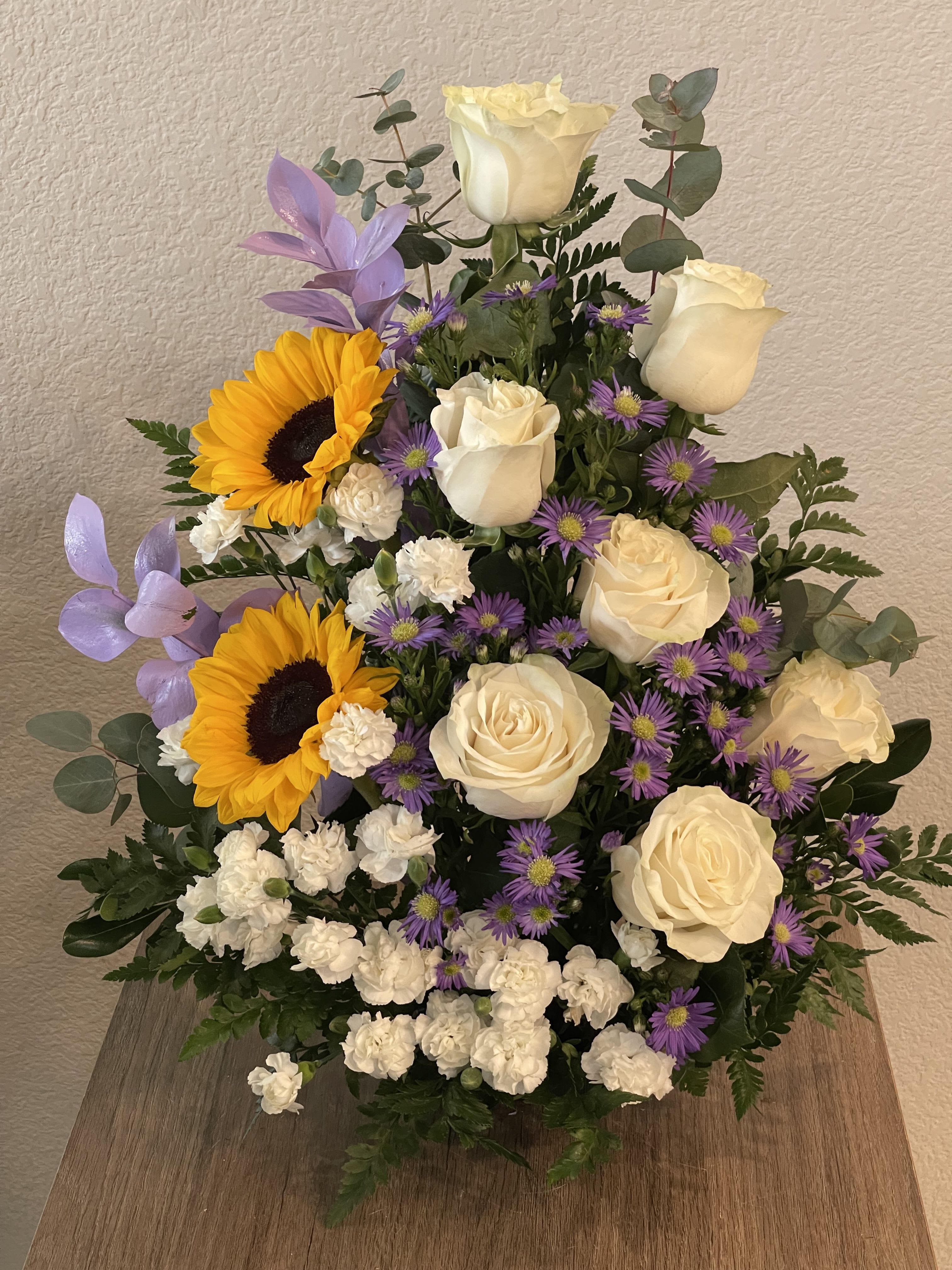 Modern Meadow Moments - Cream-white roses paired with bold yellow sunflowers create a cheerful birthday bouquet that feels like sunshine in a vase. This birthday bouquet (also perfect as a thank-you gift or a bright sympathy arrangement) mixes soft ivory roses and sunny sunflower faces with pops of purple daisy-like asters, so the person you're thinking of will feel genuinely lifted by the warm, hopeful color story.  The arrangement is full and layered with a rounded, upright silhouette for a classic yet slightly rustic garden look. White carnations and small purple asters and daisies fill the composition, adding texture and contrast against fresh green foliage; the combination of creamy petals, vivid yellow centers, and cool purple accents creates lively visual depth. Sized to sit proudly on a table or console, this bouquet brings an instantly fresh, upbeat presence to springtime celebrations, thank-you moments, or when you want to send comforting, sunny wishes.