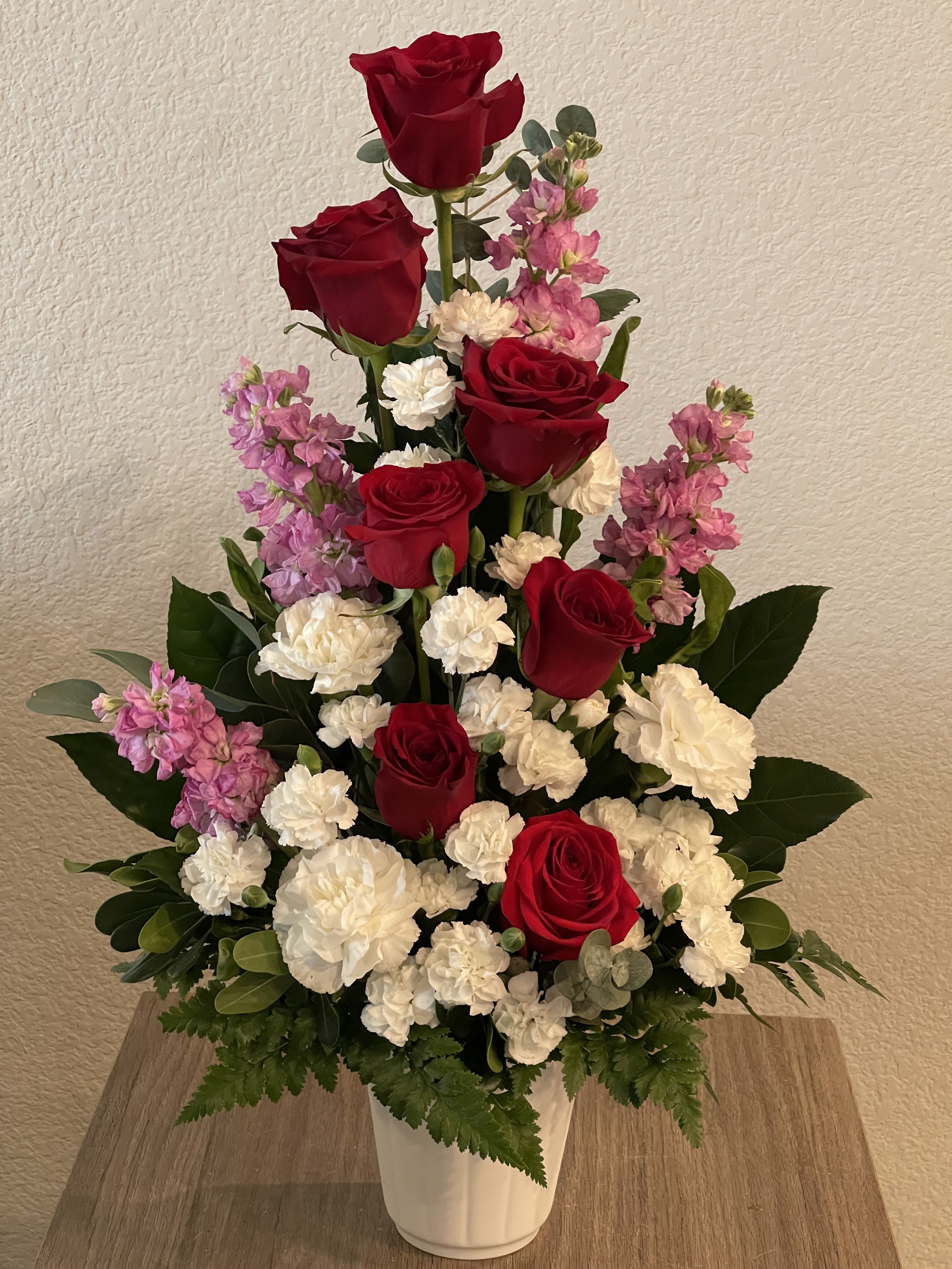 Pillar of Harmony  - Deep, velvety red roses rise through clusters of soft white carnations and pink stock in a tall, tiered bouquet-an arrangement made for anniversaries, Valentine's Day, or any moment when you want someone to feel deeply loved. The strong contrast between saturated red and gentle pink-and-white blooms reads romantic and formal, the kind of floral statement that says "you matter" to the person you're celebrating.  Closely packed roses, carnations, and stock form layered tiers framed by broad dark foliage and eucalyptus-like accents, adding texture, fullness, and a hint of green that softens the drama. The stems are presented in a white ceramic vase that gives the design a polished, timeless finish and makes it easy to display on a mantel, dining table, or entry console.  Perfect for marking a milestone, sending sympathy with dignity, or surprising the one you love, this arrangement delivers classic elegance and heartfelt sentiment. Standard care: keep in fresh water, trim stems occasionally, and place out of direct heat to extend bloom life.