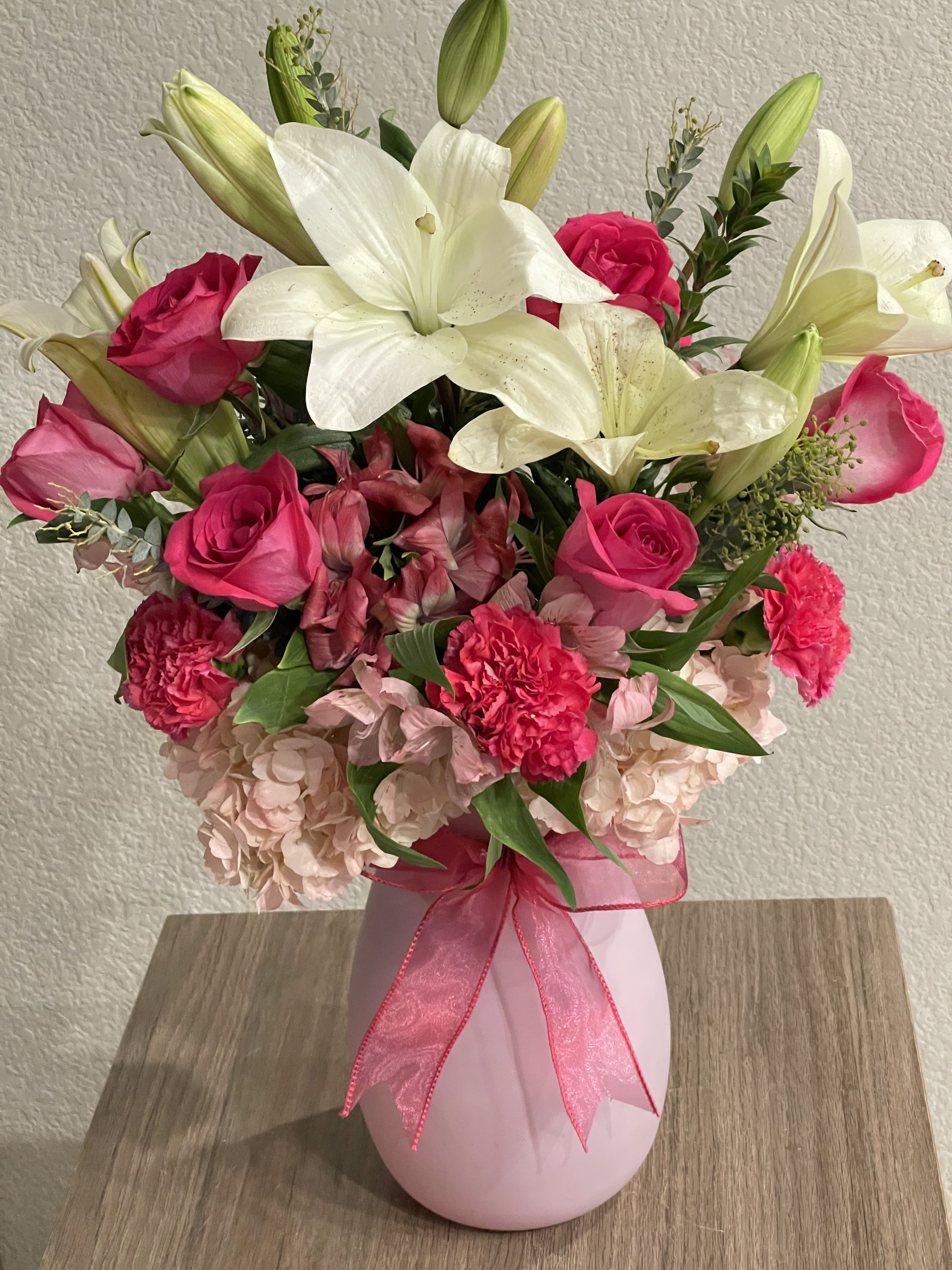 Floral Fizz - Bright white lilies rise above vivid hot-pink roses and carnations in this celebratory flower arrangement, a birthday bouquet or anniversary roses alternative that reads romantic and joyful at first glance. Soft blush hydrangeas anchor the composition while pink alstroemeria and fresh greenery add delicate texture and motion-she'll feel noticed and celebrated the moment she sees it.  Arranged in a pale pink ceramic vase and finished with a sheer pink ribbon, the bouquet balances tall lily buds for height with full, layered clusters of roses, carnations, hydrangeas, and alstroemeria for volume and contrast. The color story-pink, white, and green-feels feminine and bright without being over-the-top, making it equally right for Mother's Day, a heartfelt thank-you, or to mark a special anniversary.  This is the kind of arrangement that reads as both polished gift and warm sentiment for the person you're thinking of; the sturdy ceramic vase arrives ready to display. Standard care-fresh water and a cool spot away from direct sun-will help the lilies and blooms open beautifully over several days.