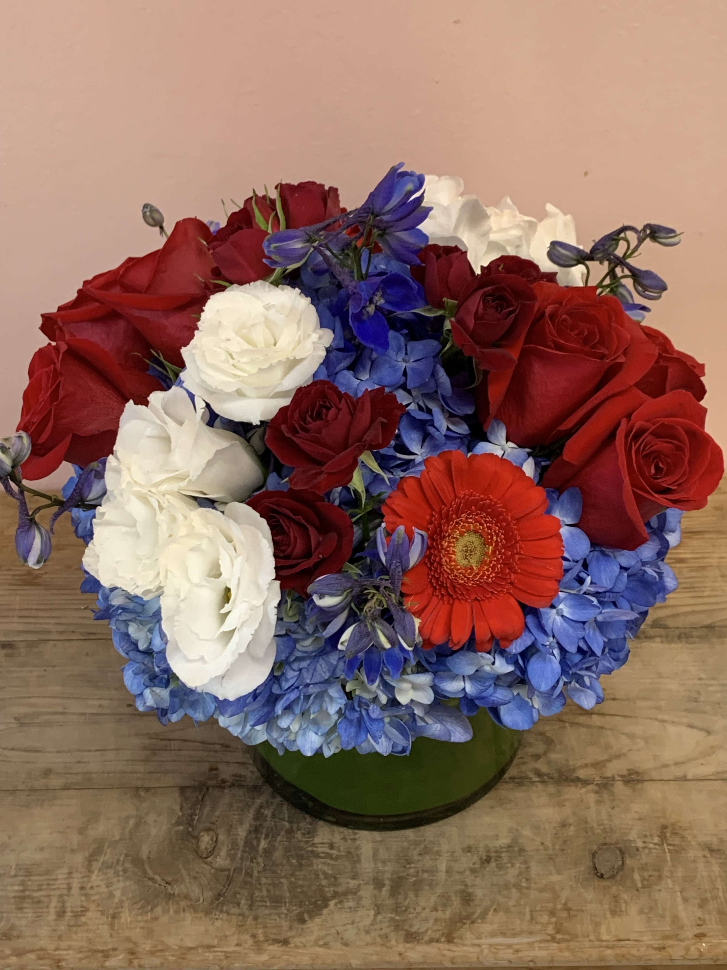 Red and white roses with blue hydrangeas and an orange gerbera daisy in a green vase