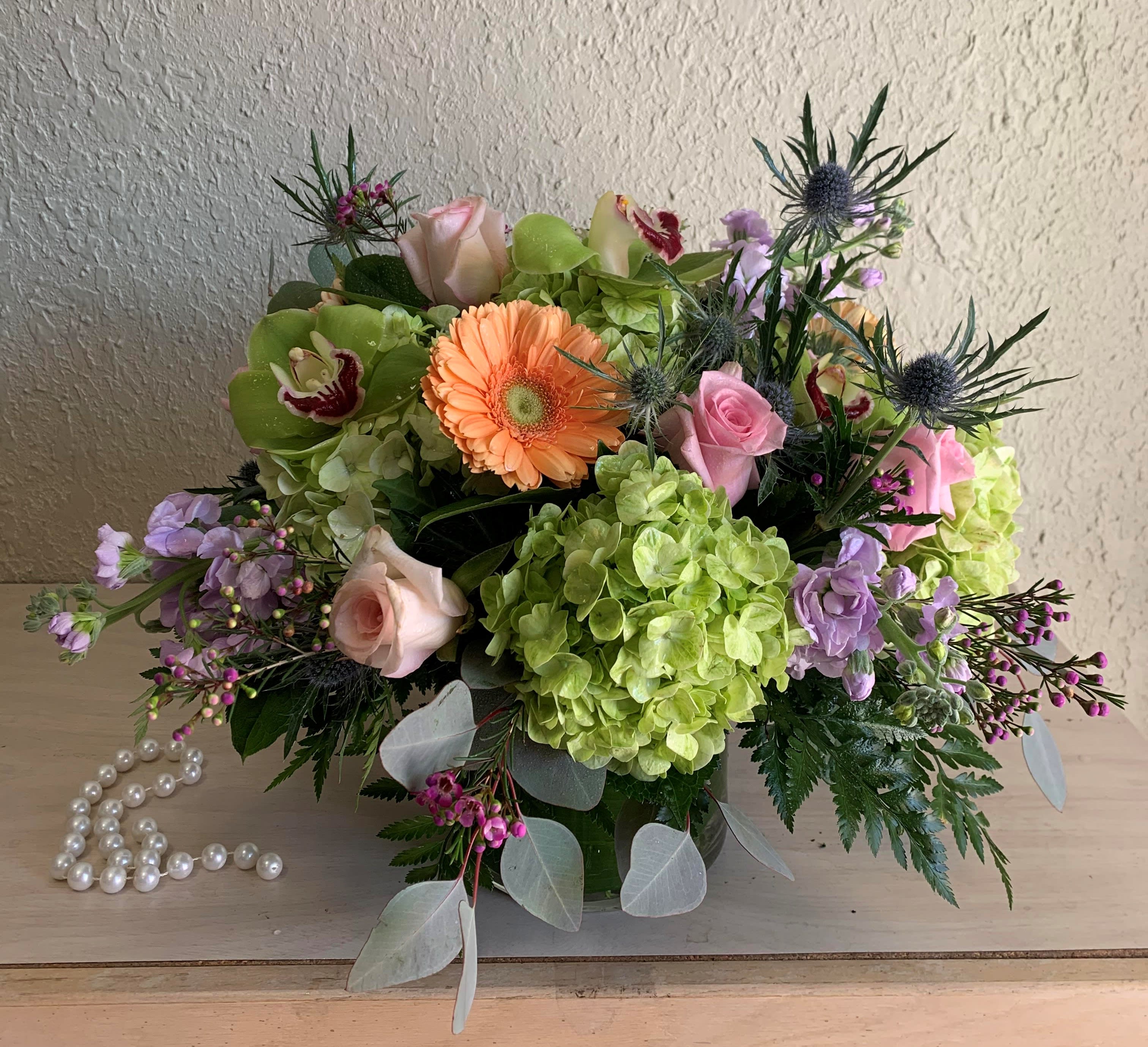 Low arrangement of green hydrangeas, peach gerbera, pink roses, orchids in a glass vase beside pearl beads