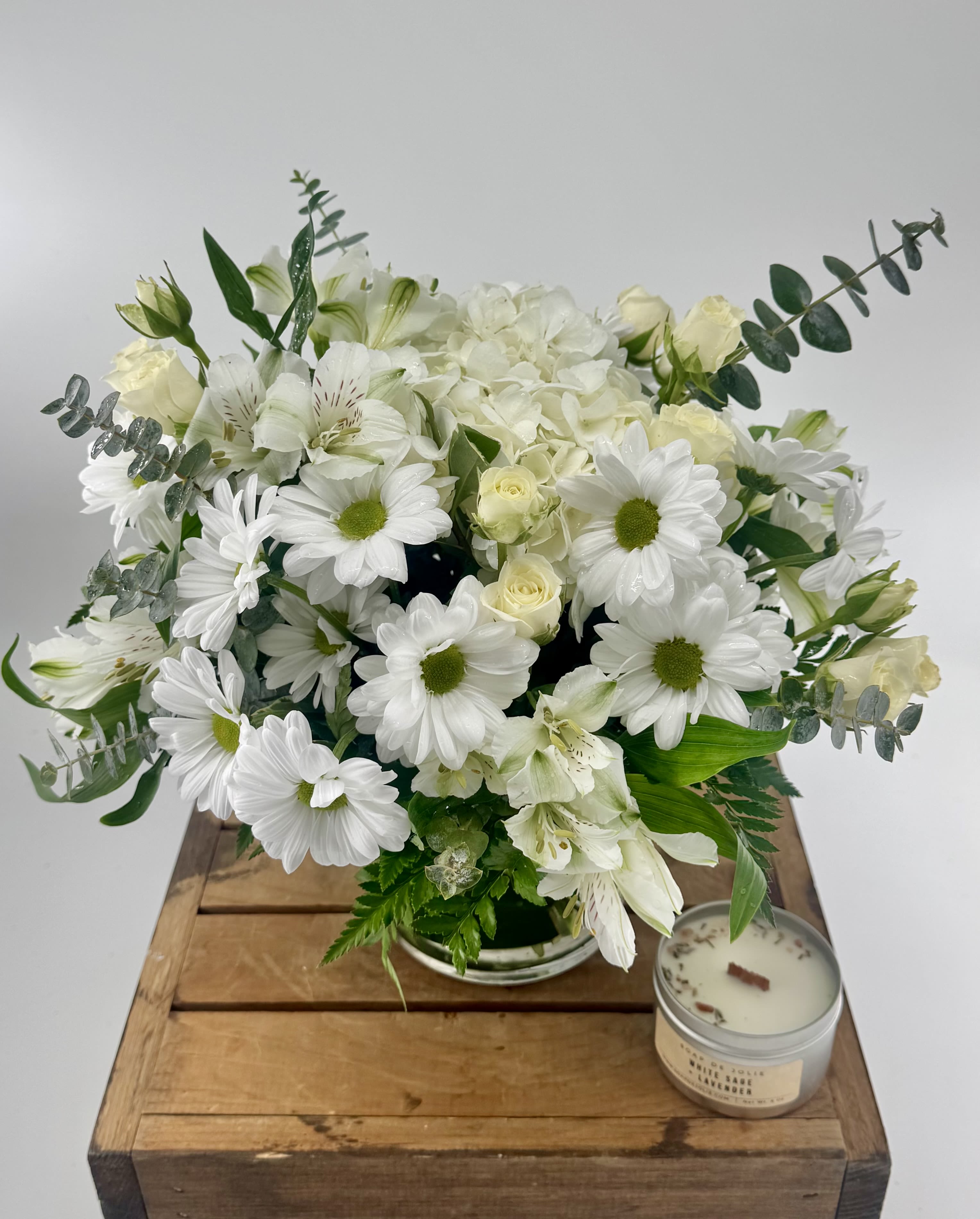 White daisies, roses, and hydrangeas in a glass vase beside a tin candle on a wooden crate.