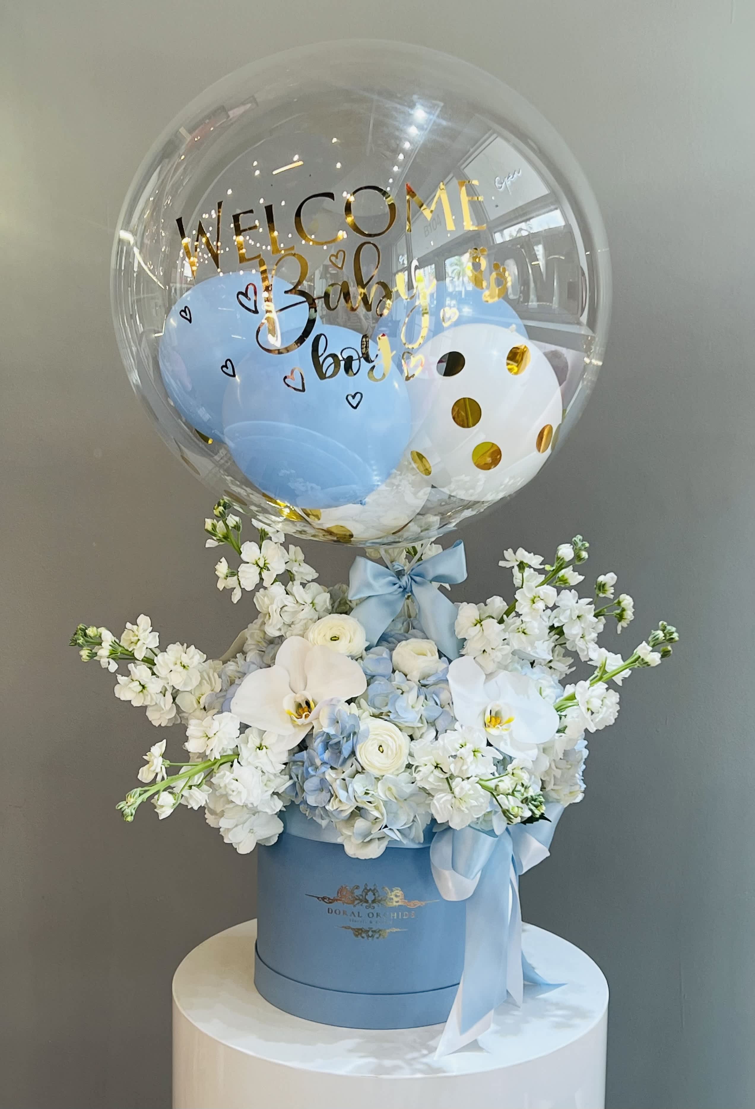 Welcome Baby Boy! ™ - A joyful welcome arrangement, this design pairs soft blue hydrangeas, elegant white orchids, and delicate ranunculus with airy white blooms for a fresh, serene look. Topped with a festive balloon and tied with satin blue ribbons, it’s a charming and celebratory piece—perfect for greeting a precious baby boy.