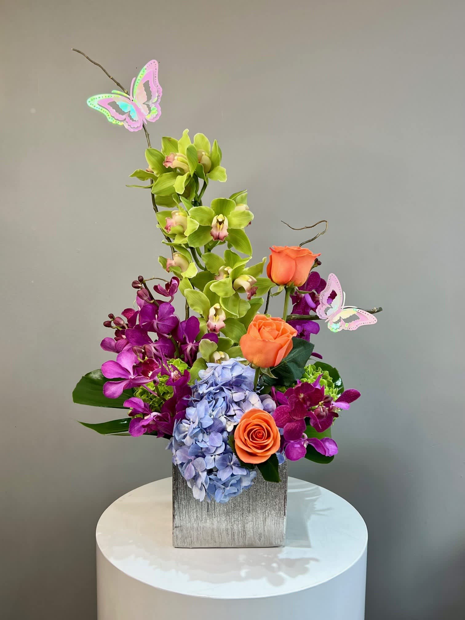 Tall arrangement of green and purple orchids, orange roses, and blue hydrangea in a square vase with butterfly picks
