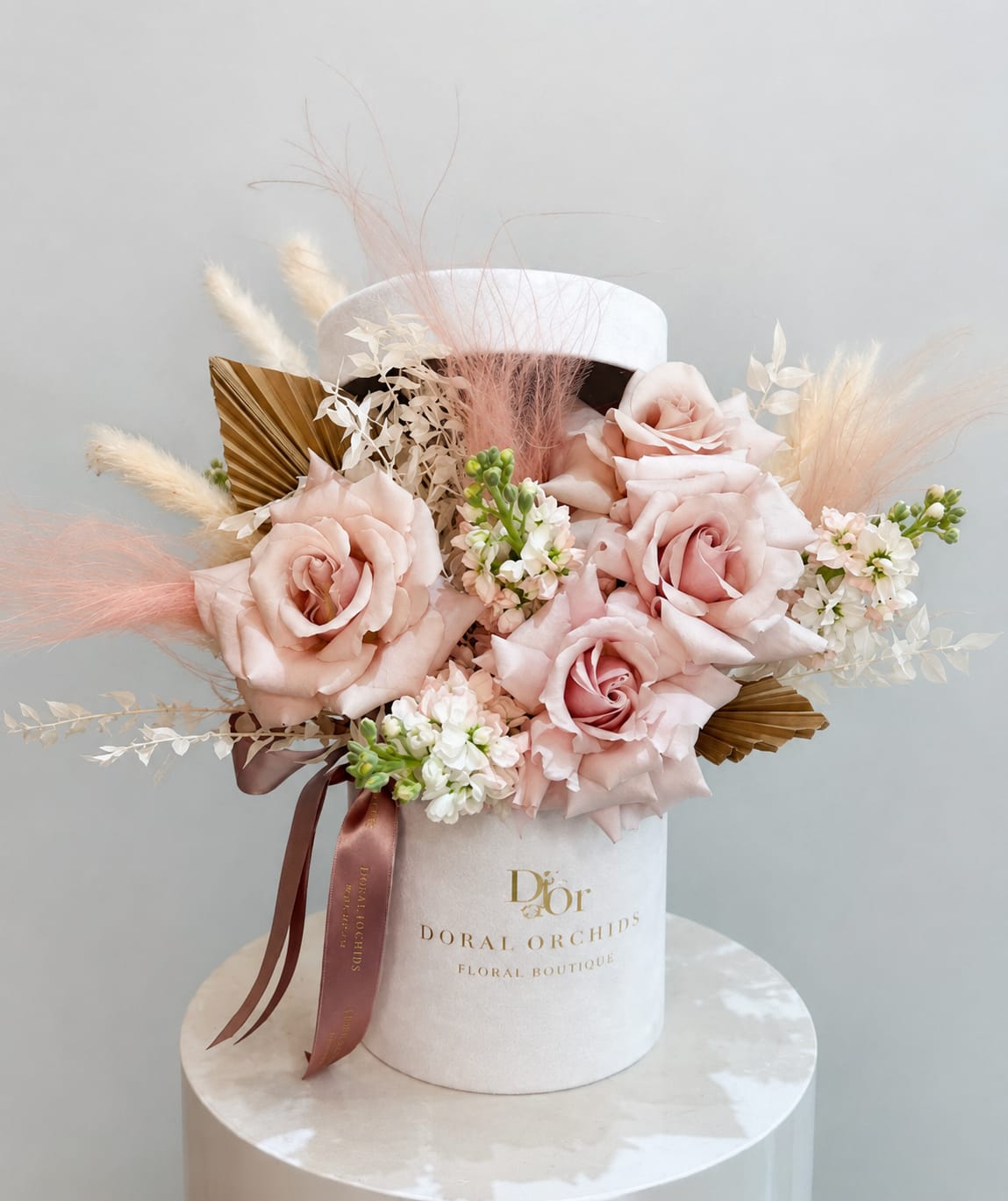 Sahara ™ - Velvety blush roses take center stage in this arrangement, softened by airy pampas grass, golden palms, and delicate white blooms. Presented in a suede hatbox with satin ribbons, it radiates romantic charm and refined elegance.