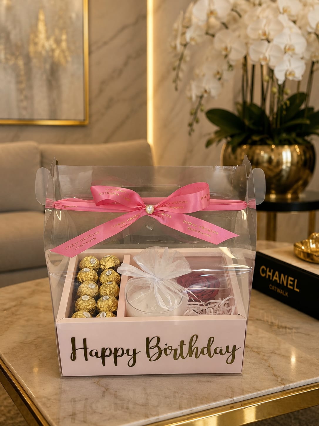 Blush Birthday Luxe Gift Box ™ - A single red rose set against soft pink and white accents makes a quietly luxurious birthday gift-perfect as a birthday box for a romantic partner or someone you want to indulge. The Blush Birthday Luxe Gift Box reads polished and celebratory: a clear presentation box tied with a pink ribbon and a bold "Happy Birthday" message announces the occasion while keeping the rose as the emotional centerpiece.  Inside, the arrangement pairs one long-stemmed red rose with white shredded filler and gold-wrapped chocolate truffles, plus a small glass candle for a warm, personal touch. The color story blends pink, white and metallic gold for a refined look; the clear box lets the textures-velvety petals, shiny wrappers, and the soft glow of the candle-show through for immediate visual impact.  This box is designed to make the recipient feel cherished and seen on their birthday-romantic enough for anniversaries, thoughtful enough for a close friend who likes a touch of luxury. Ready to gift as pictured, it arrives assembled and ribboned; light the candle gently and keep the rose in water if you transfer it to a vase to extend its life.
