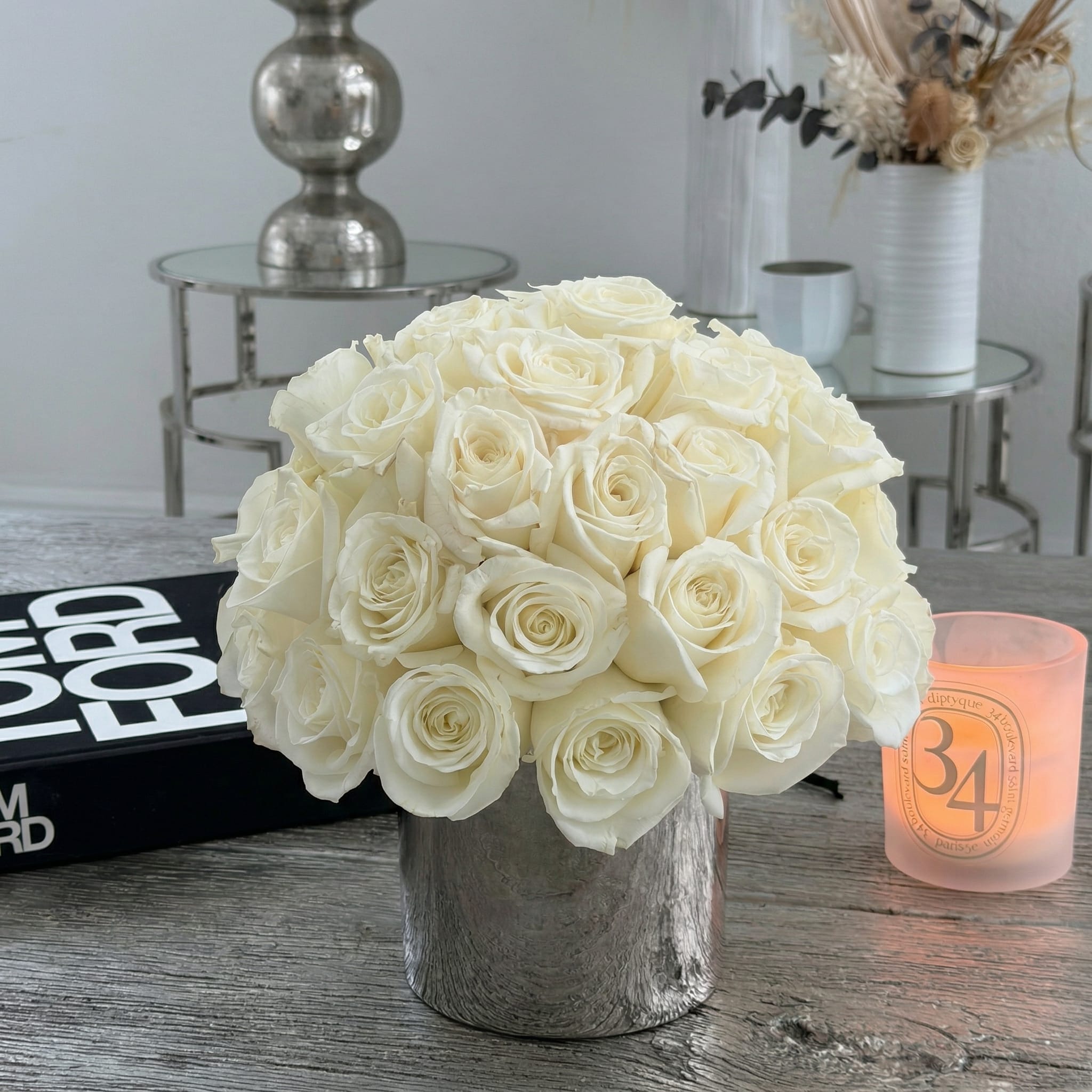 Compact arrangement of white roses in a silver vase on a table with a candle and book.