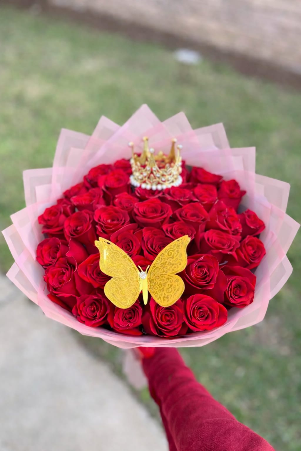 Handheld bouquet of red roses wrapped in pink paper with a gold butterfly and crown decoration.