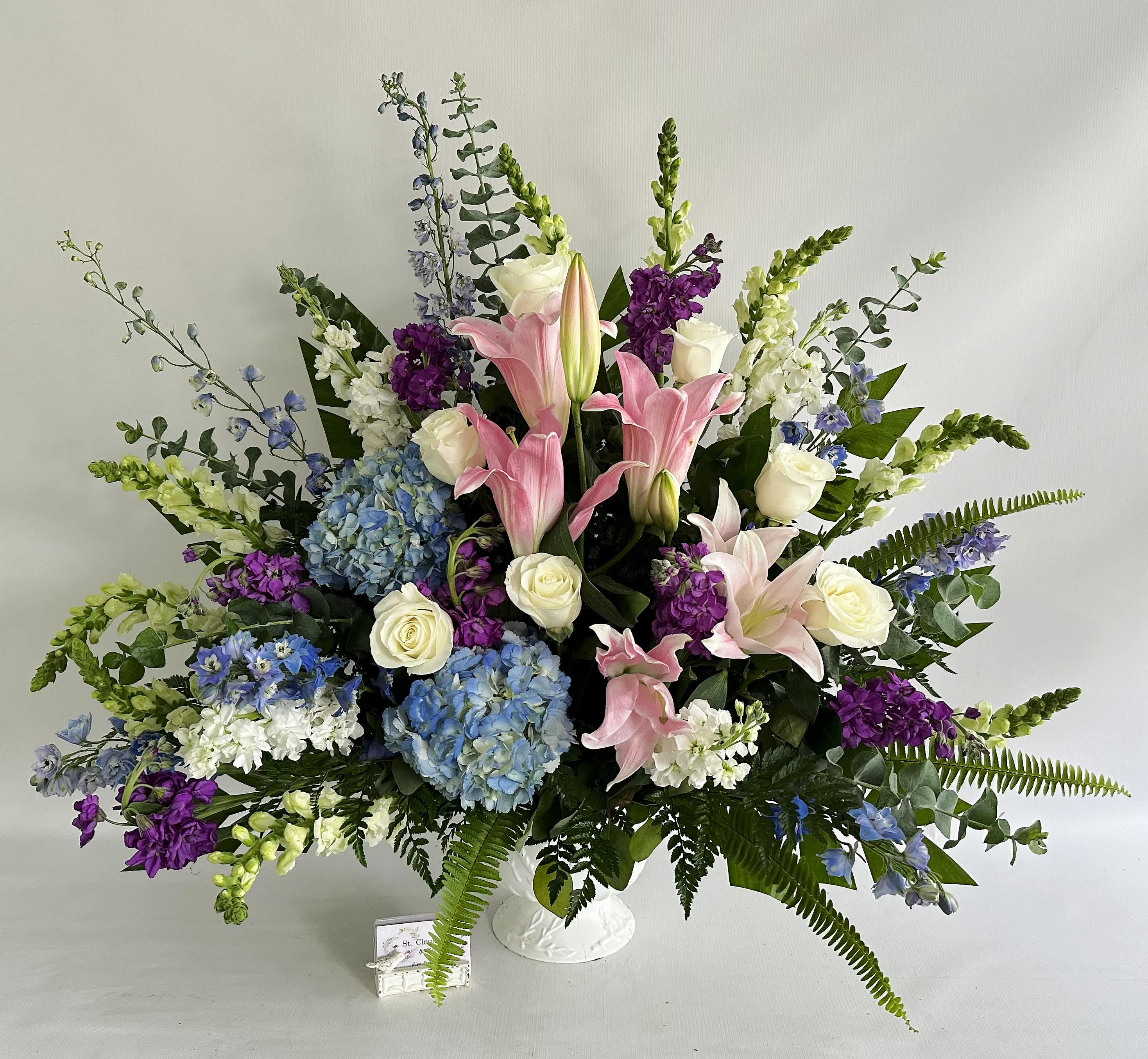 Large bouquet of pink lilies, white roses, blue hydrangeas, and purple flowers in a white vase.