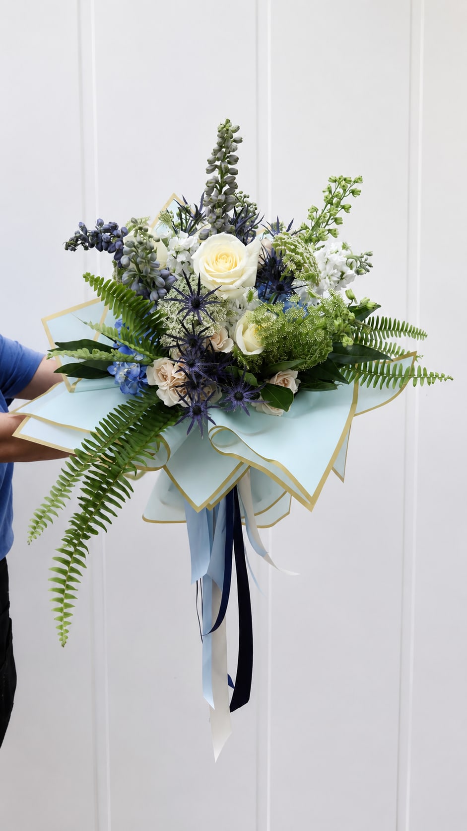 Handheld bouquet of white roses, blue flowers, and fern fronds wrapped in pale blue paper