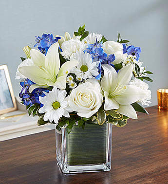 White lilies, roses, daisies, and blue flowers in a square glass vase