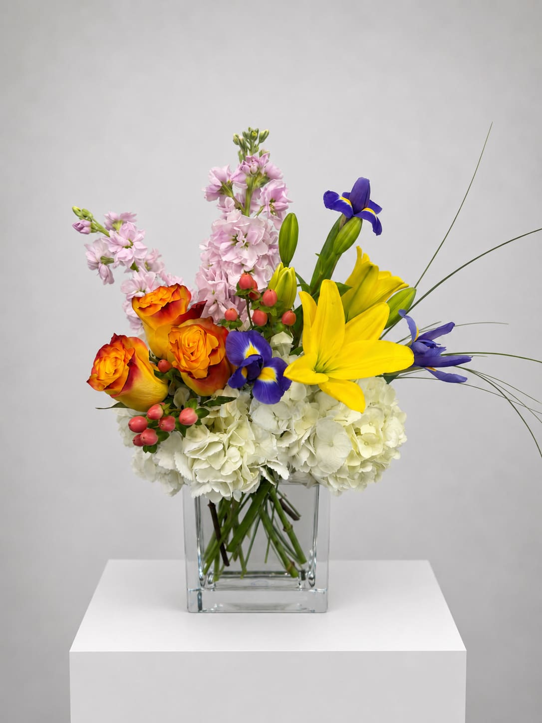 Mixed bouquet in a clear square vase with yellow lilies, orange roses, and blue irises