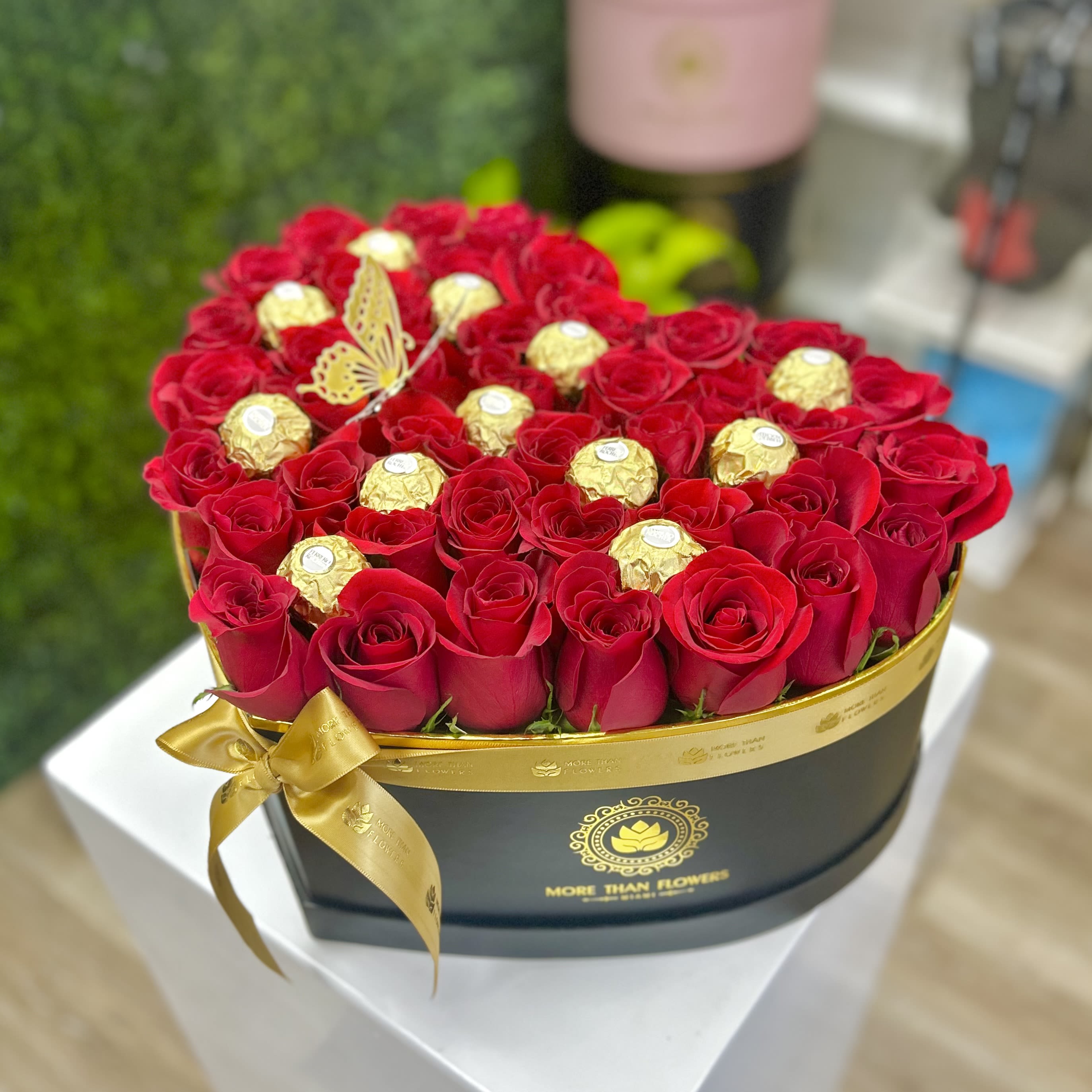 Heart-shaped black box of red roses with gold-wrapped chocolates and a gold ribbon bow.