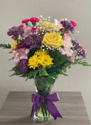 Mixed bouquet of yellow, pink, and purple flowers in a glass vase with a purple ribbon