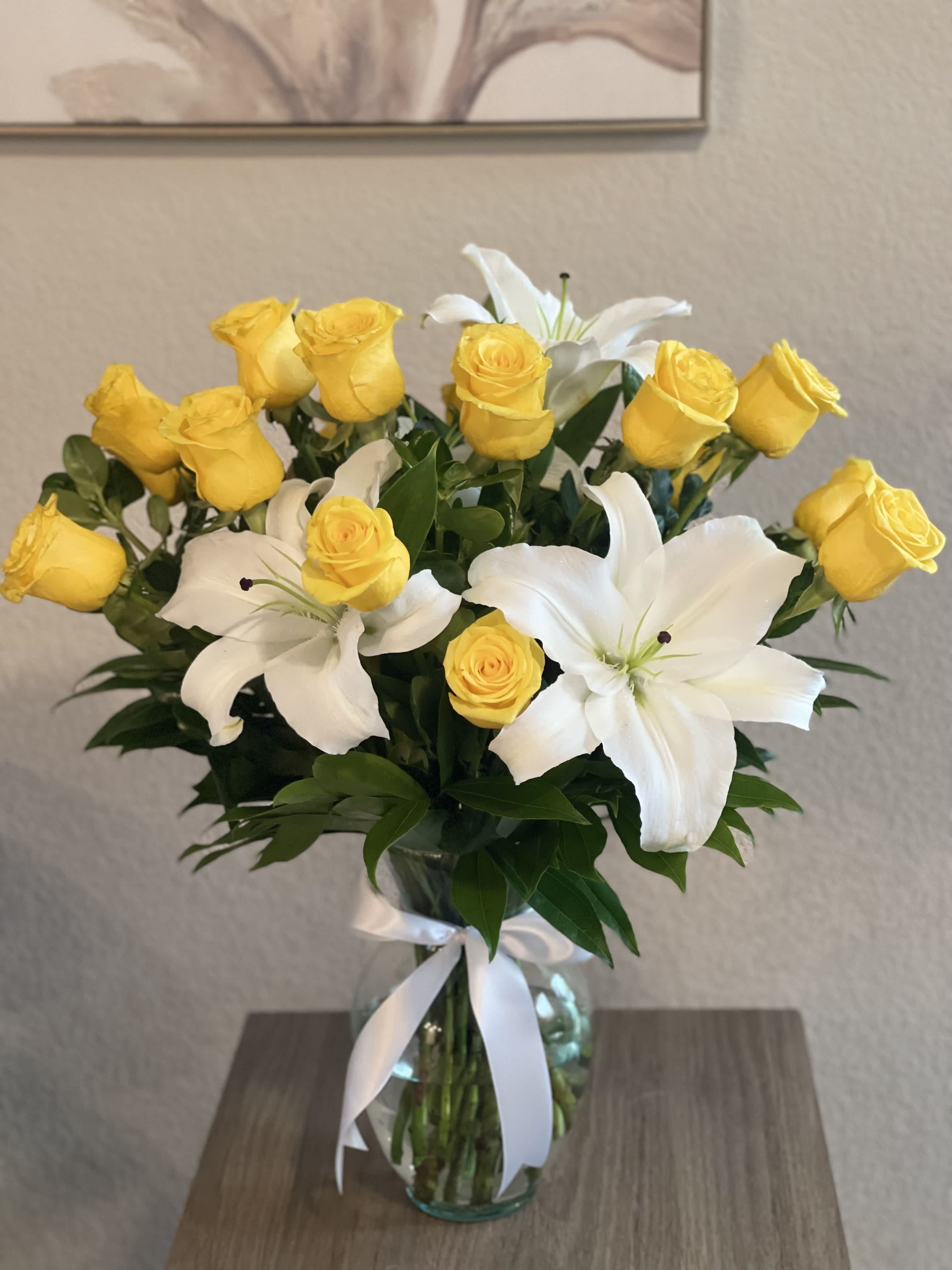 Yellow roses and white lilies arranged in a glass vase with a white ribbon.