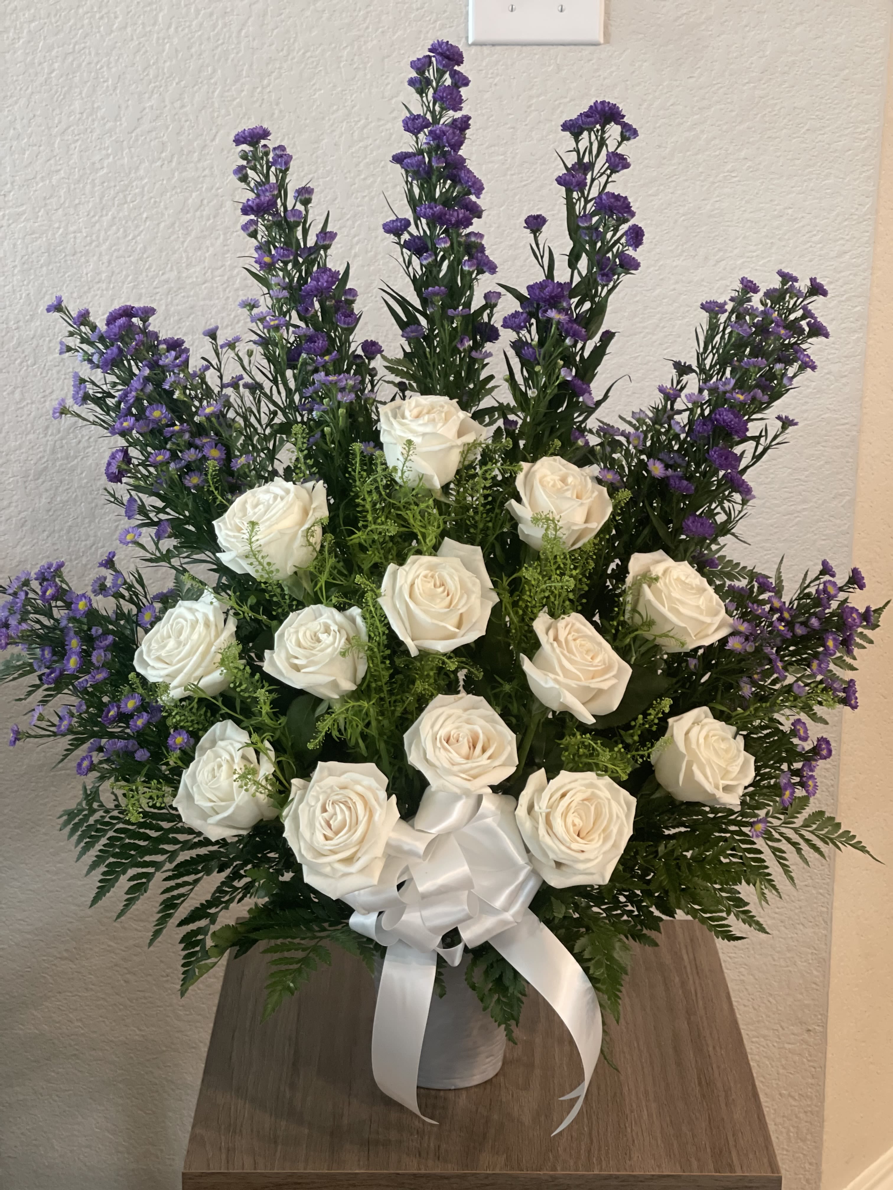 White roses and purple filler flowers in a white vase with a ribbon.